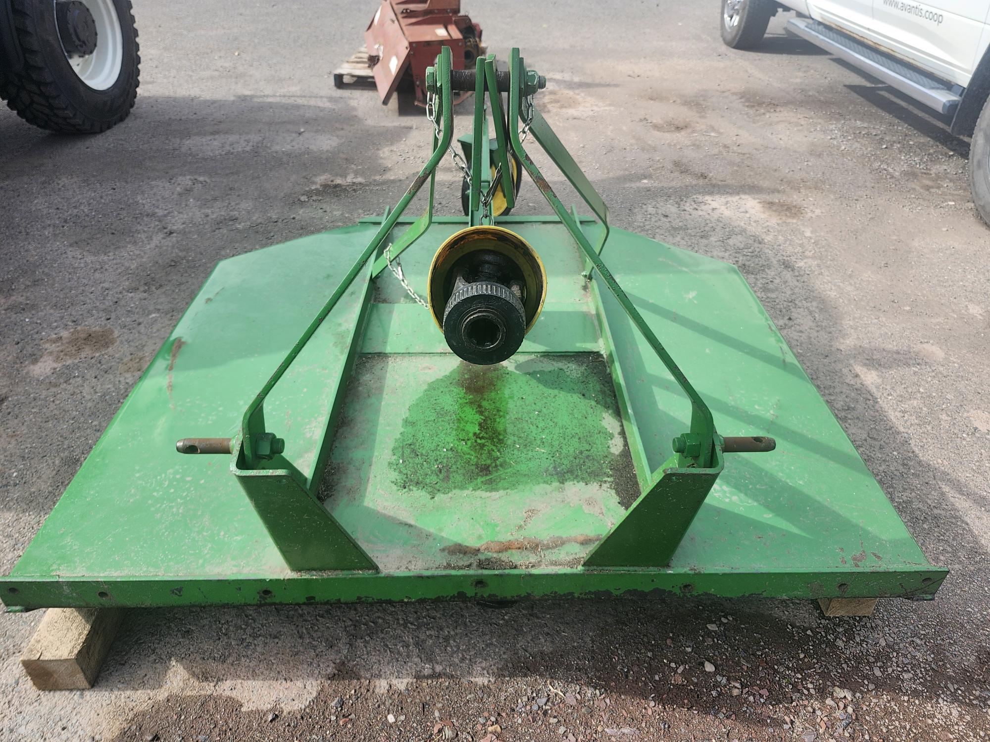 Un accessoire agricole vert de forme carrée, peut-être une tondeuse ou une débroussailleuse, repose sur le sol. Il est composé d'un élément circulaire central et de barres métalliques s'étendant jusqu'aux bords. À proximité se trouvent les roues d'un véhicule et d'un équipement agricole.