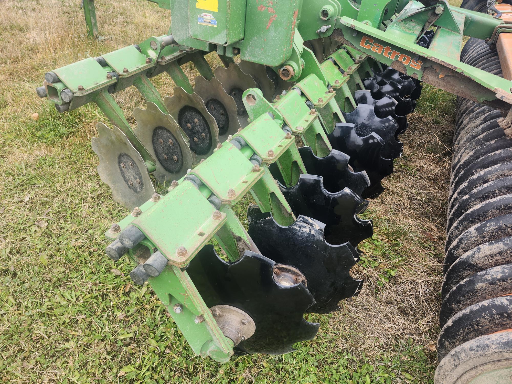 Gros plan d'une herse à disques agricole verte dotée de lames circulaires tranchantes conçues pour le travail du sol. L'équipement est positionné sur un terrain herbeux et présente une certaine usure due à son utilisation.