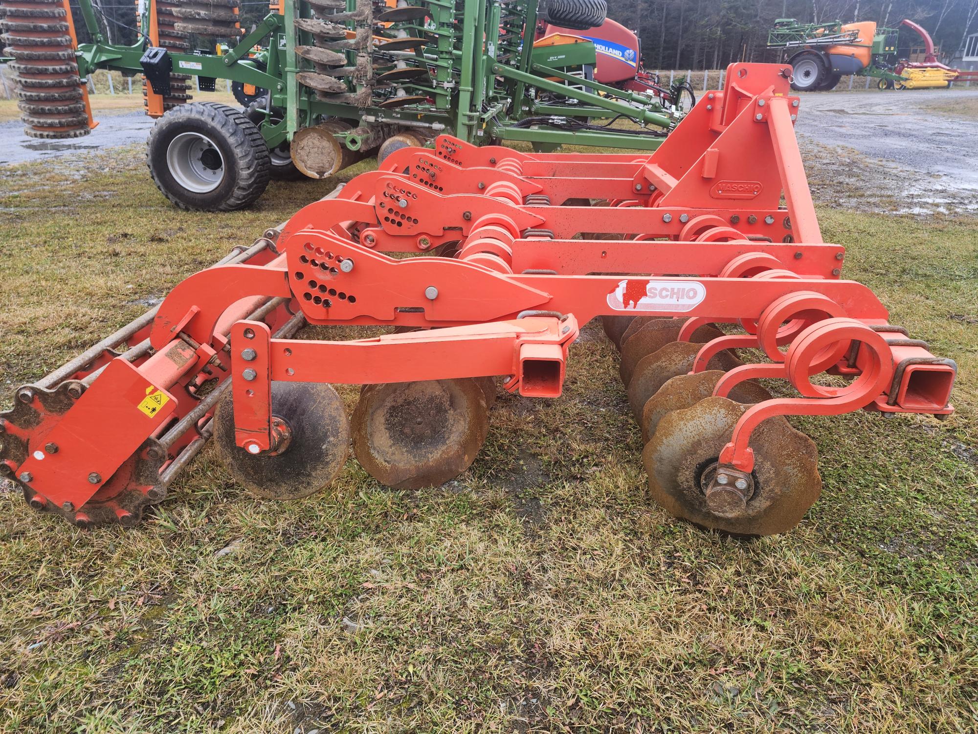 Une herse à disques agricole rouge à plusieurs lames rondes est garée sur un sol herbeux. La machine est conçue pour labourer le sol, avec des roues visibles en arrière-plan.