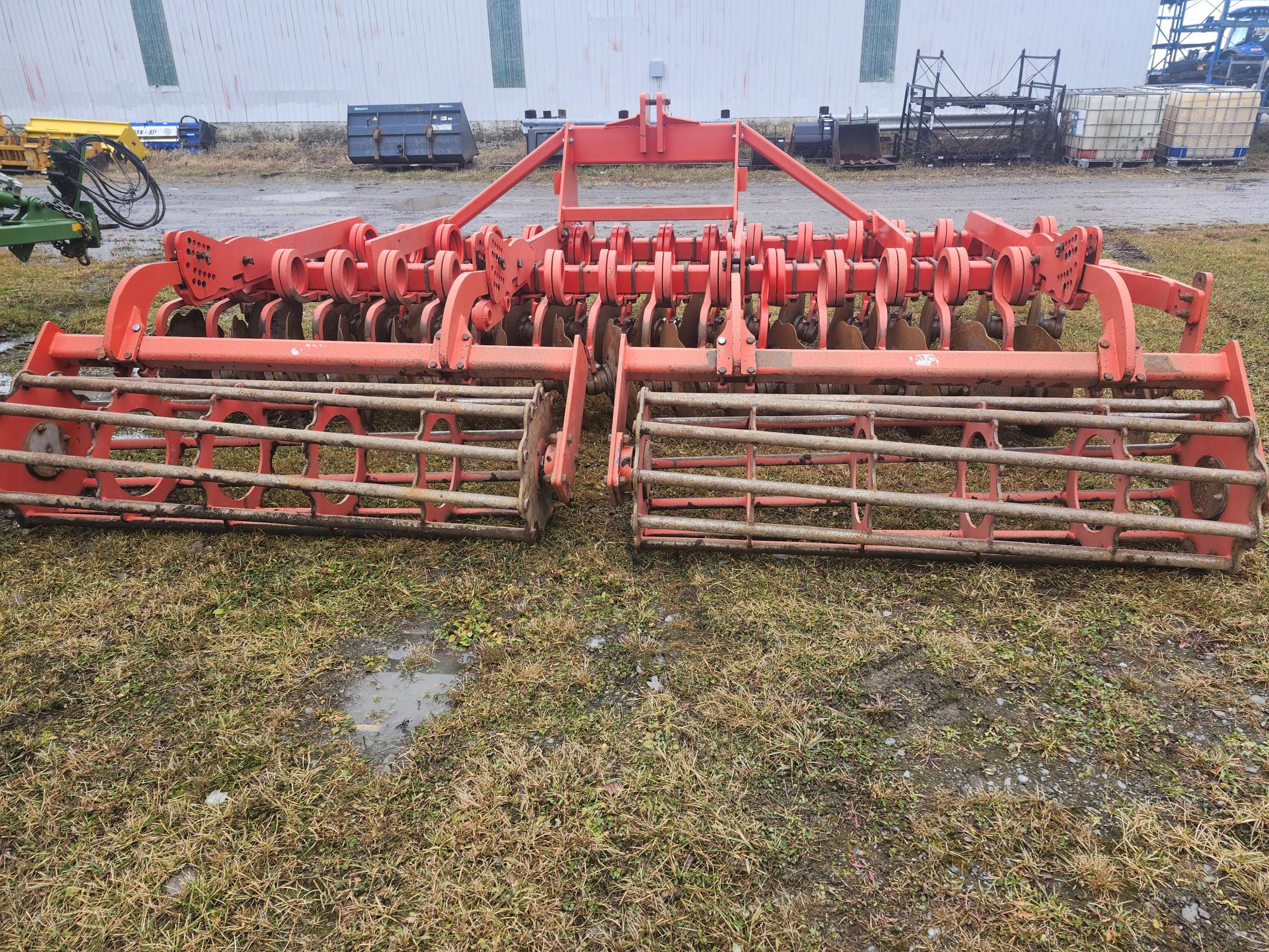 Un outil agricole rouge avec plusieurs disques et rouleaux, posé sur une parcelle herbeuse. Il semble patiné, avec un entrepôt et d'autres équipements agricoles en arrière-plan.