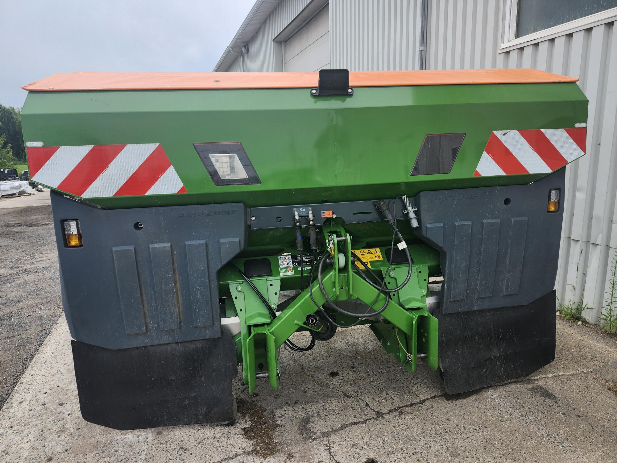 Une machine agricole verte et grise est garée devant un bâtiment métallique. Son toit est orange vif avec des rayures diagonales rouges et blanches à l'arrière. Divers composants mécaniques sont visibles en dessous.