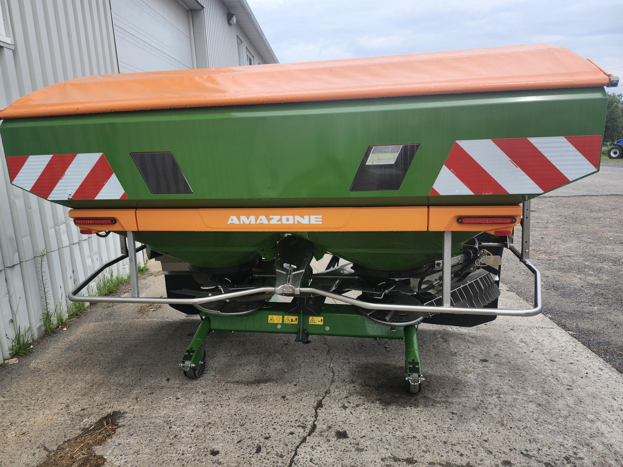 Une machine agricole verte et orange avec des bandes réfléchissantes rouges et blanches, étiquetée « Amazone », est garée à l'extérieur d'un bâtiment sur une surface en béton. La machine est utilisée pour épandre des matériaux dans les champs.