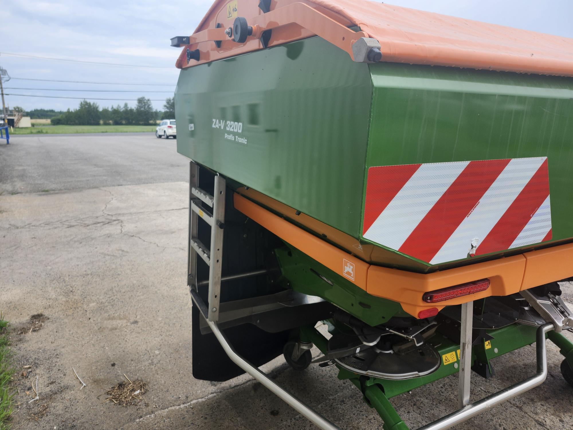 Une machine agricole verte et orange portant le numéro de modèle « ZAV 3200 » est garée sur une surface pavée. Elle porte un panneau d'avertissement rayé rouge et blanc à l'arrière. L'arrière-plan montre un champ ouvert et un ciel nuageux.