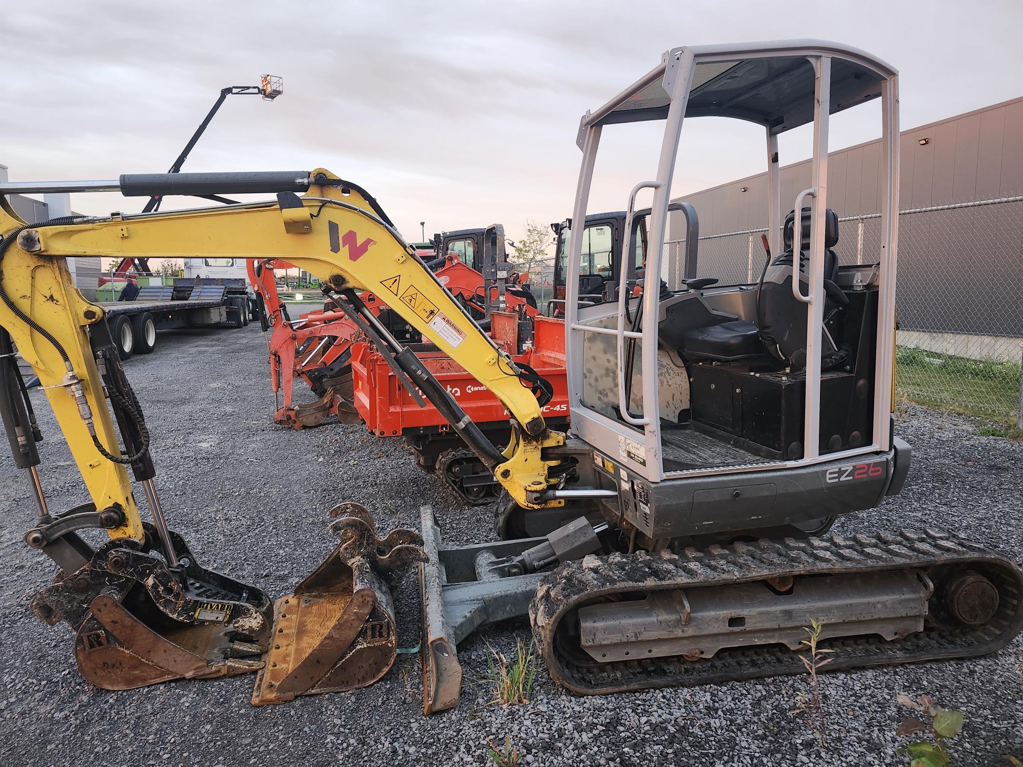 Une petite excavatrice jaune et grise avec une cabine ouverte est garée sur du gravier. Derrière elle se trouvent deux machines orange et un bâtiment avec une clôture. Le ciel est couvert.