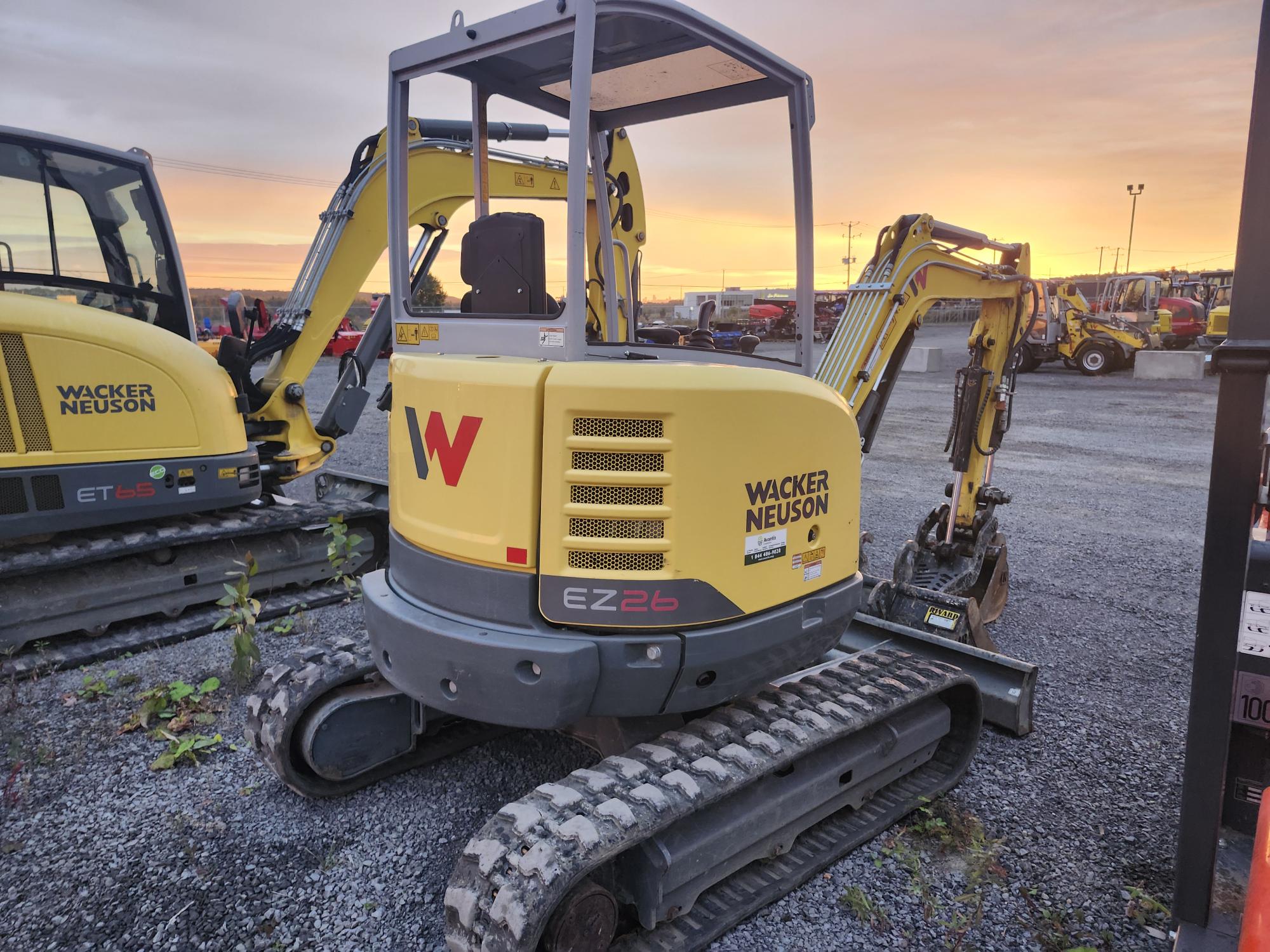 Une mini-pelle jaune Wacker Neuson EZ26 est garée sur du gravier dans un parc de matériel de construction au coucher du soleil. La machine est équipée de chenilles et d'un petit bras de creusement. Une autre machine similaire est visible à proximité.