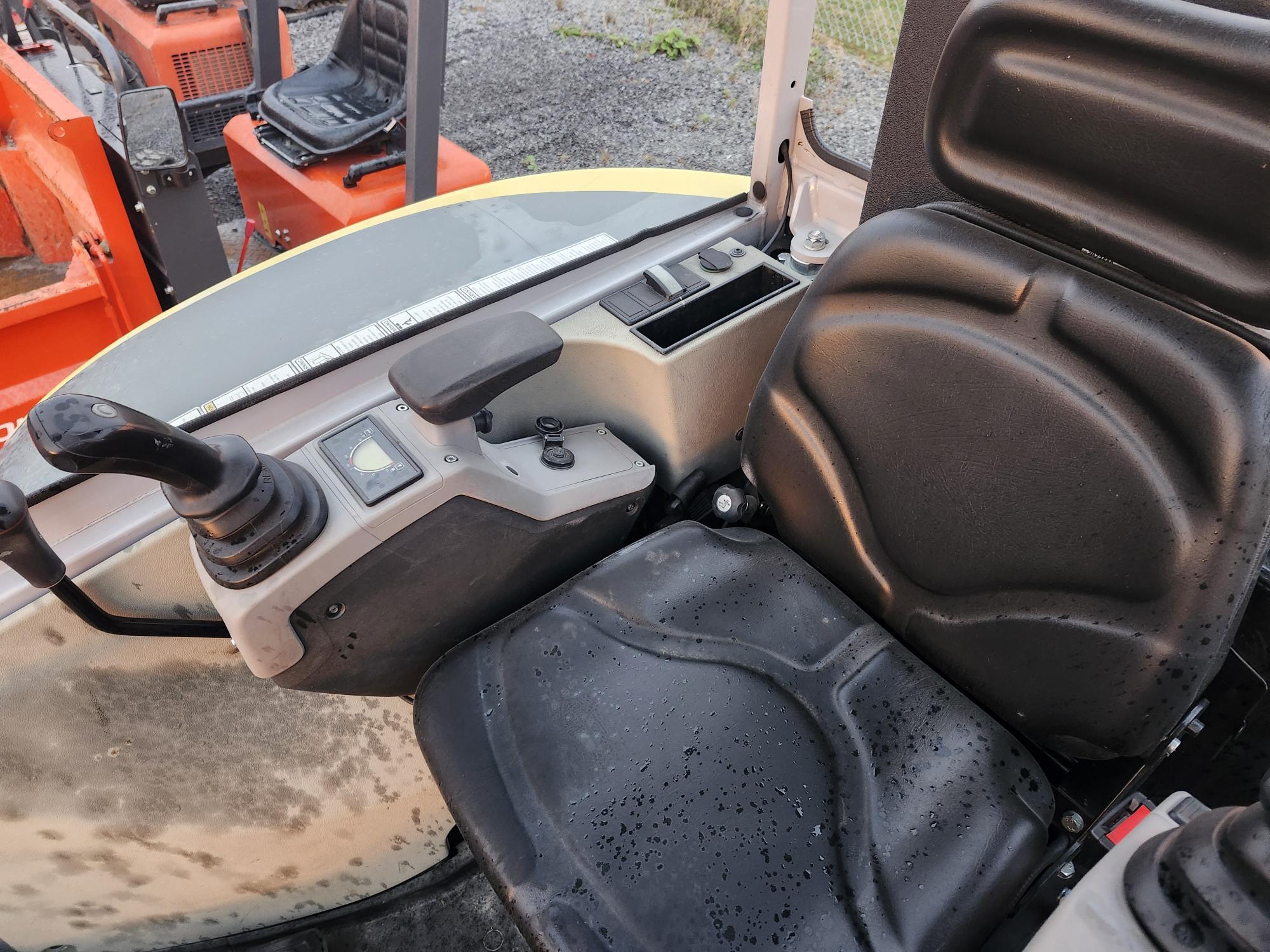 Gros plan sur un siège noir avec des gouttes d'eau dans le cockpit d'un engin de chantier. Le cockpit est doté d'un levier de commande et d'un tableau de bord avec diverses commandes et affichages. Des engins de chantier orange sont visibles à l'extérieur.