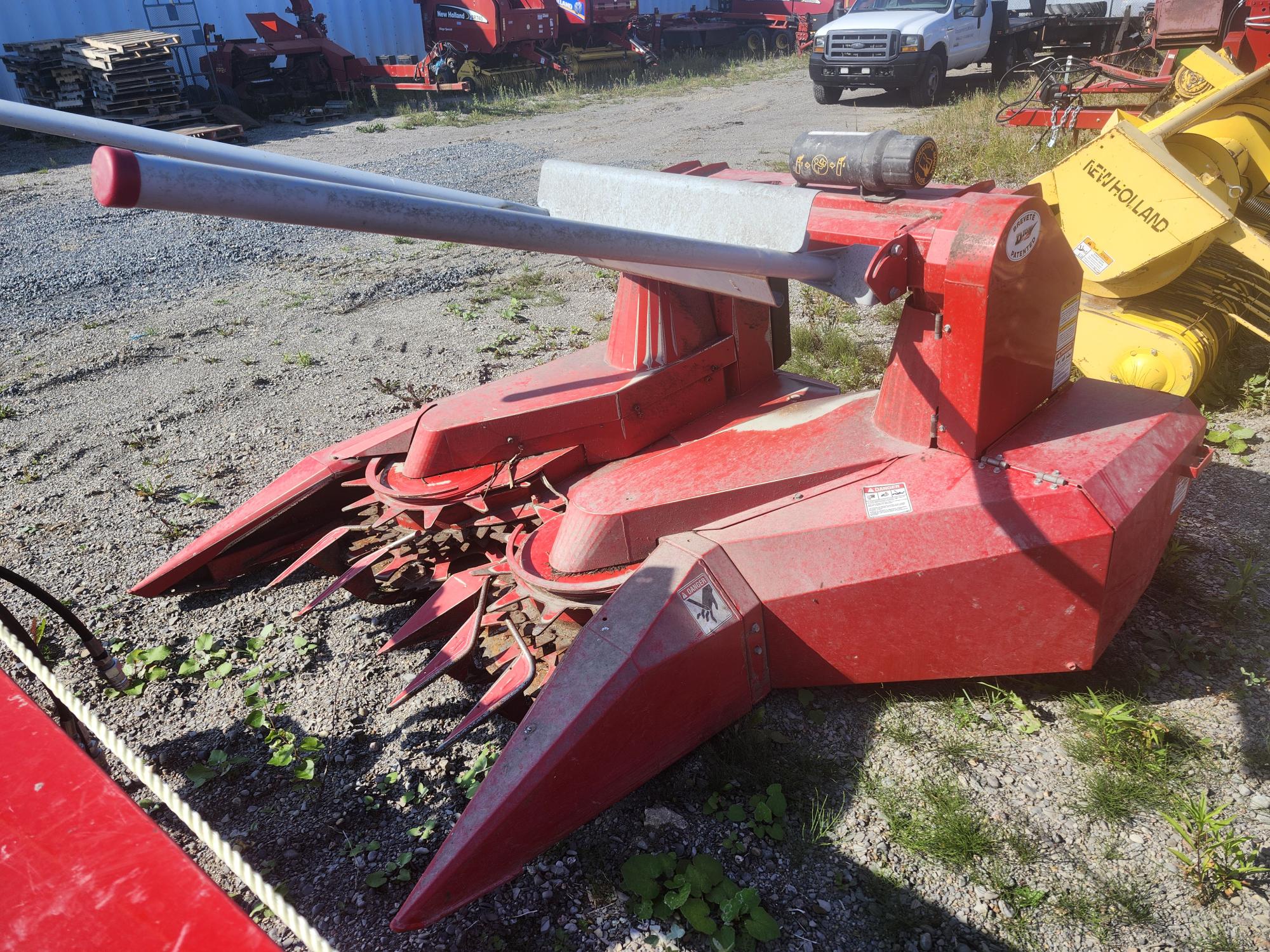 Un accessoire de machine agricole rouge sur une surface en gravier, doté de plusieurs dents métalliques et de disques rotatifs pour la coupe. Autour se trouvent d'autres machines et un camion blanc garé.