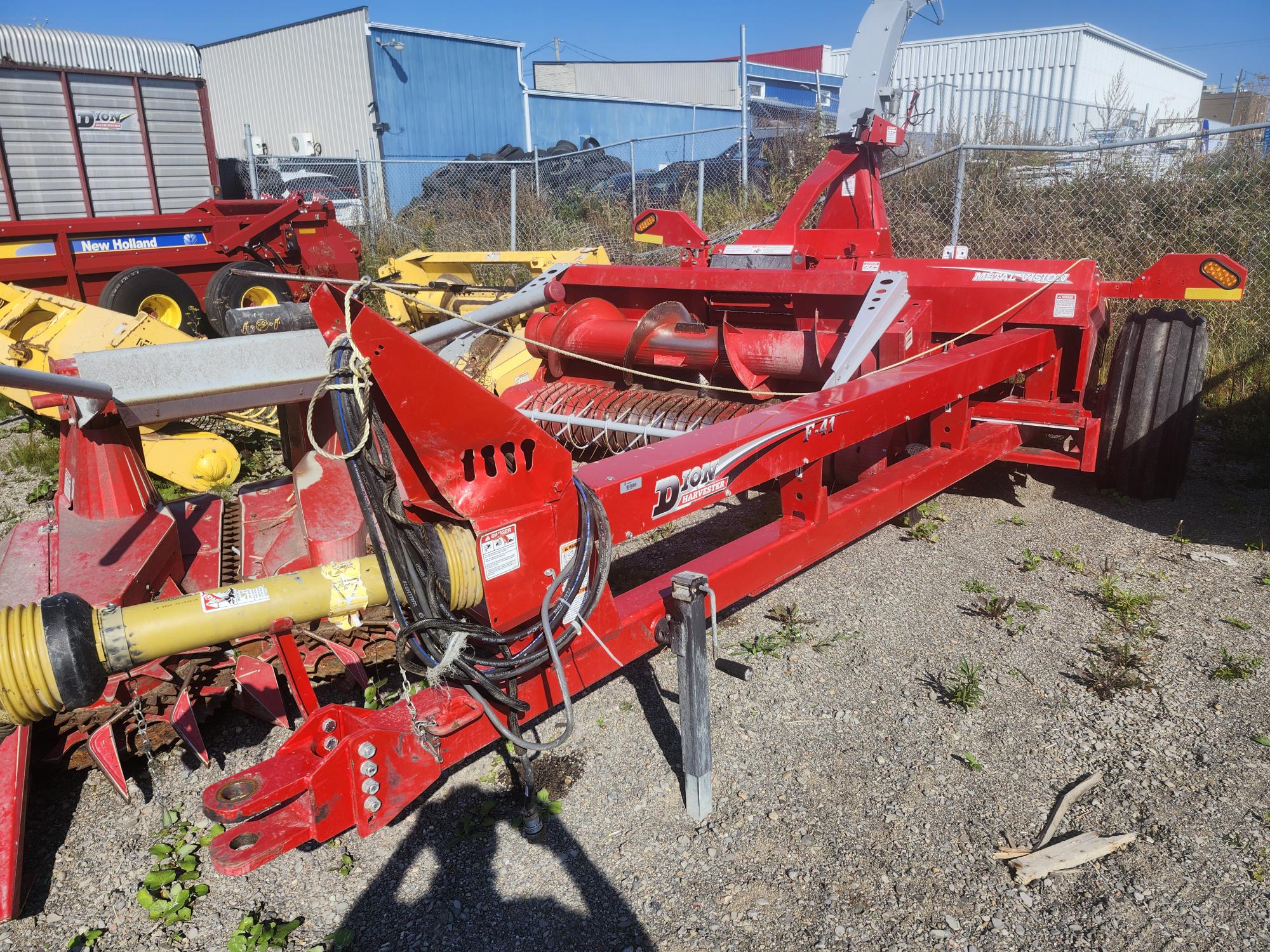 Une machine agricole rouge avec un accessoire jaune est garée sur une surface en gravier. Elle est entourée d'autres équipements agricoles, avec des bâtiments industriels et une clôture grillagée en arrière-plan.