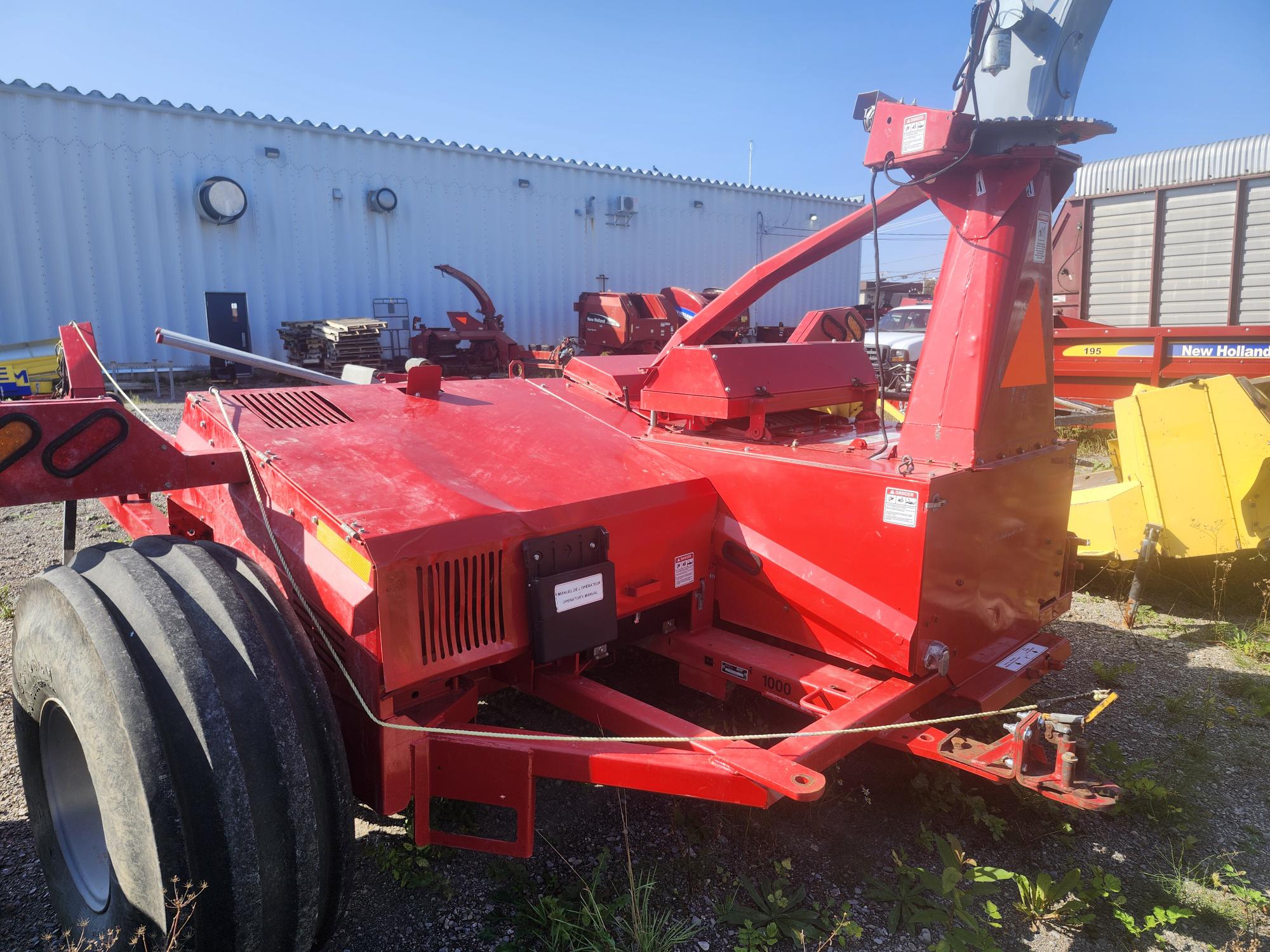 Une grande machine agricole rouge avec des roues jumelées et des pièces métalliques pointues est placée parmi d'autres machines à l'extérieur. Un bâtiment industriel blanc avec des ouvertures rondes se trouve à l'arrière-plan sous un ciel bleu clair.