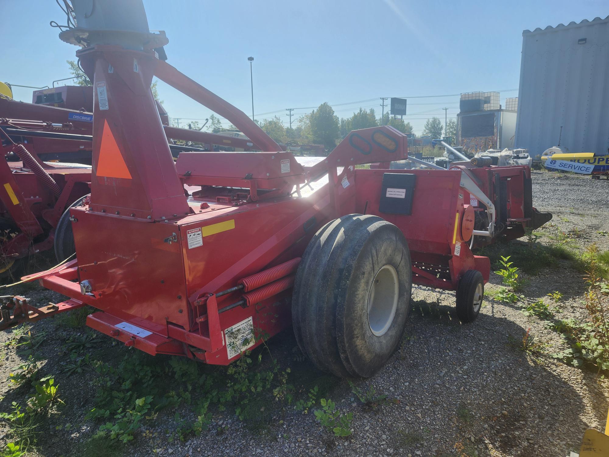 Machines agricoles rouges garées sur du gravier, entourées de plantes vertes. Un panneau de sécurité triangulaire lumineux est visible et un ciel bleu clair apparaît en arrière-plan.