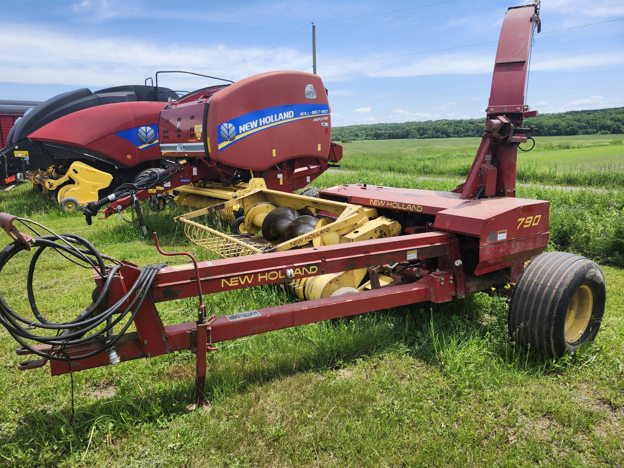 Une ensileuse New Holland rouge et jaune est garée sur un champ herbeux avec une presse à balles rouge en arrière-plan. La scène comprend des champs verts luxuriants et un ciel bleu.