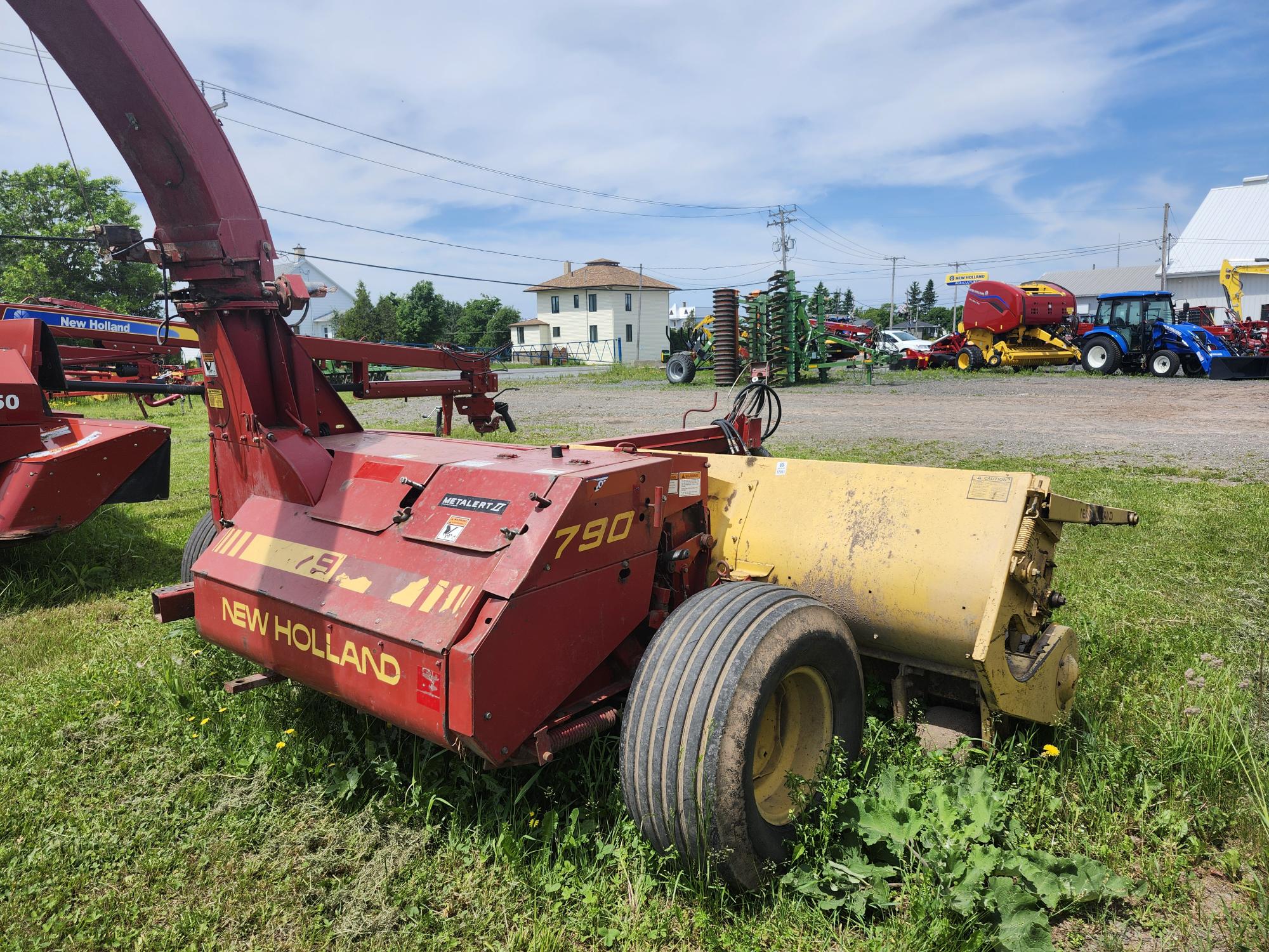 Une ensileuse New Holland rouge et jaune portant le numéro de modèle 790 est posée sur l'herbe dans un champ ouvert. D'autres équipements agricoles sont visibles en arrière-plan sous un ciel partiellement nuageux.