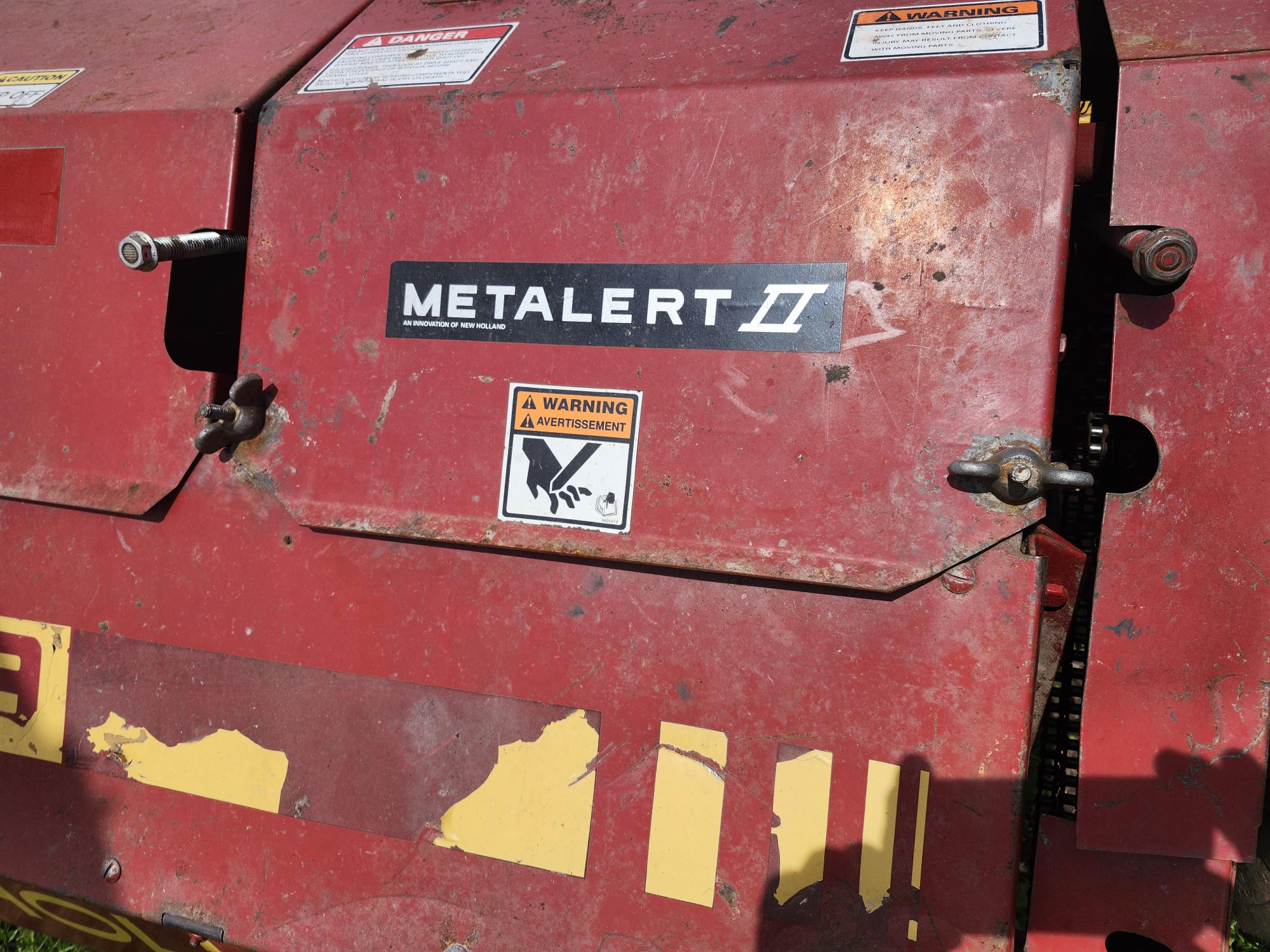 Gros plan d'une machine agricole rouge portant une étiquette indiquant « METALERT II » et un autocollant d'avertissement représentant un danger pour les mains. La surface de la machine présente des signes d'usure et des marquages jaunes décolorés.