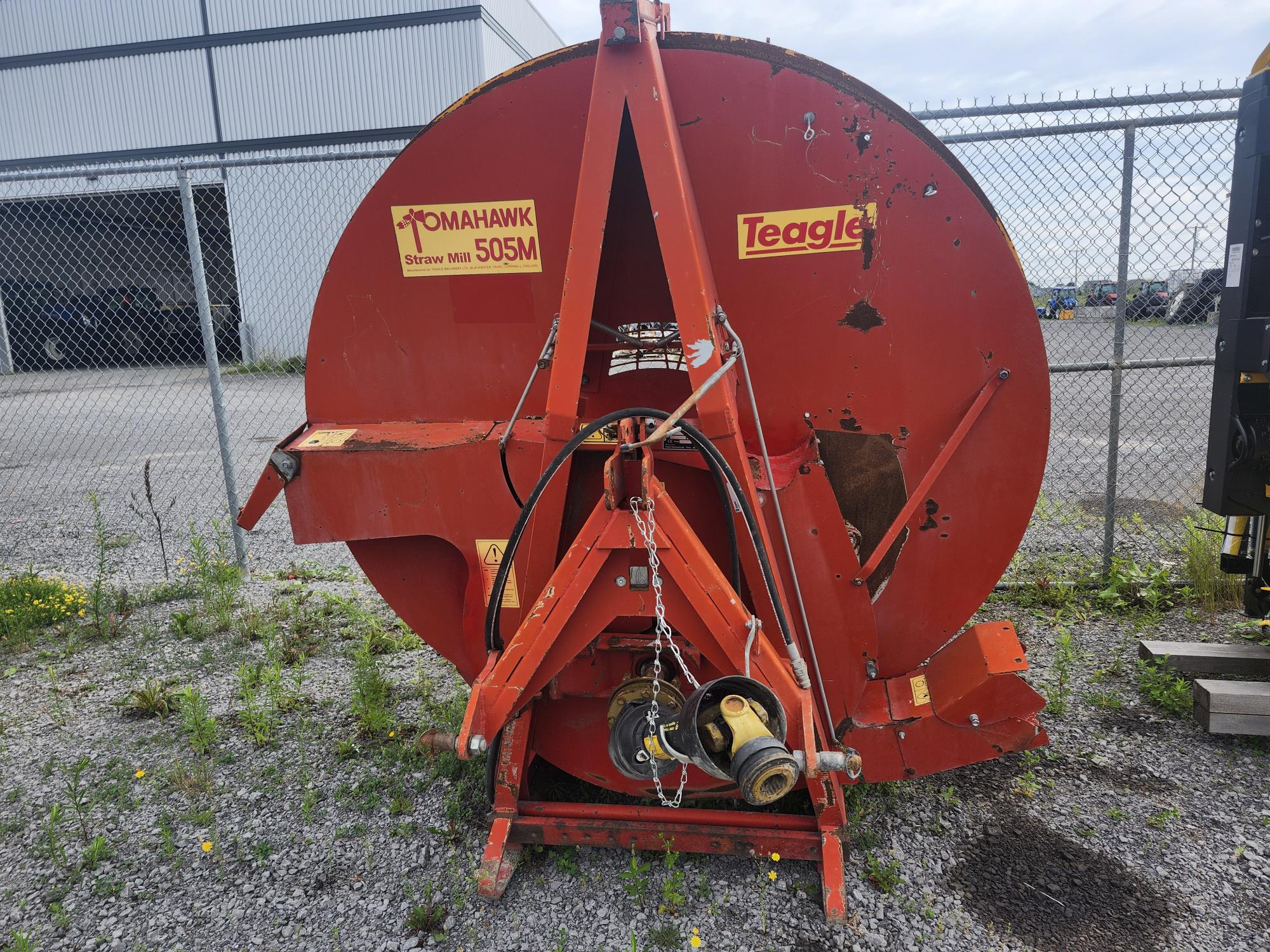 Moulin à paille Red Teagle Tomahawk 505M posé sur du gravier à côté d'une clôture grillagée, avec des bâtiments industriels en arrière-plan. La machine est patinée et on peut voir de l'herbe pousser à travers le gravier.