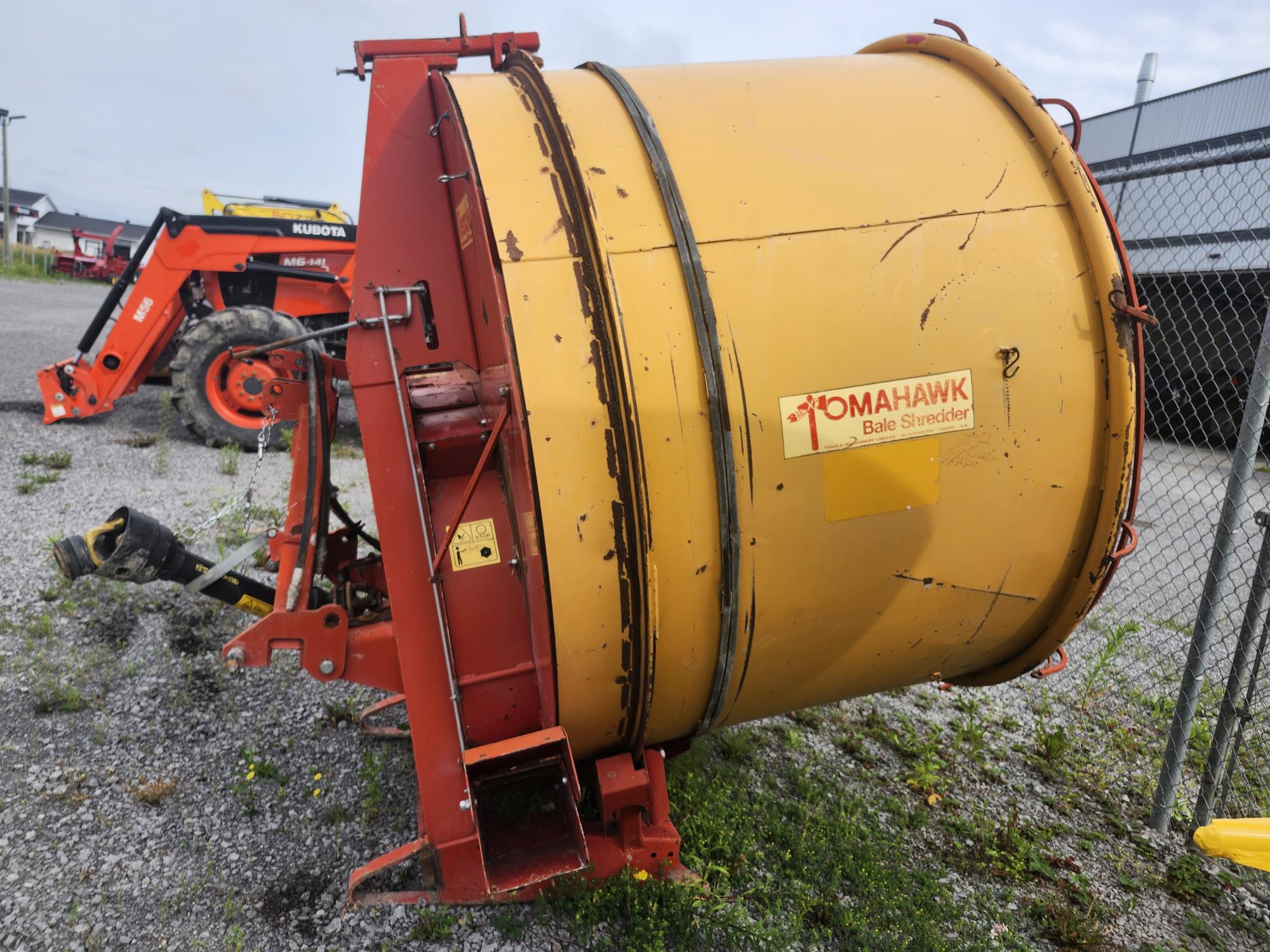 Une grande machine agricole jaune et rouge étiquetée « Tomahawk Bale Shredder » est garée sur une surface en gravier près d'une clôture grillagée. Un tracteur rouge est visible en arrière-plan.