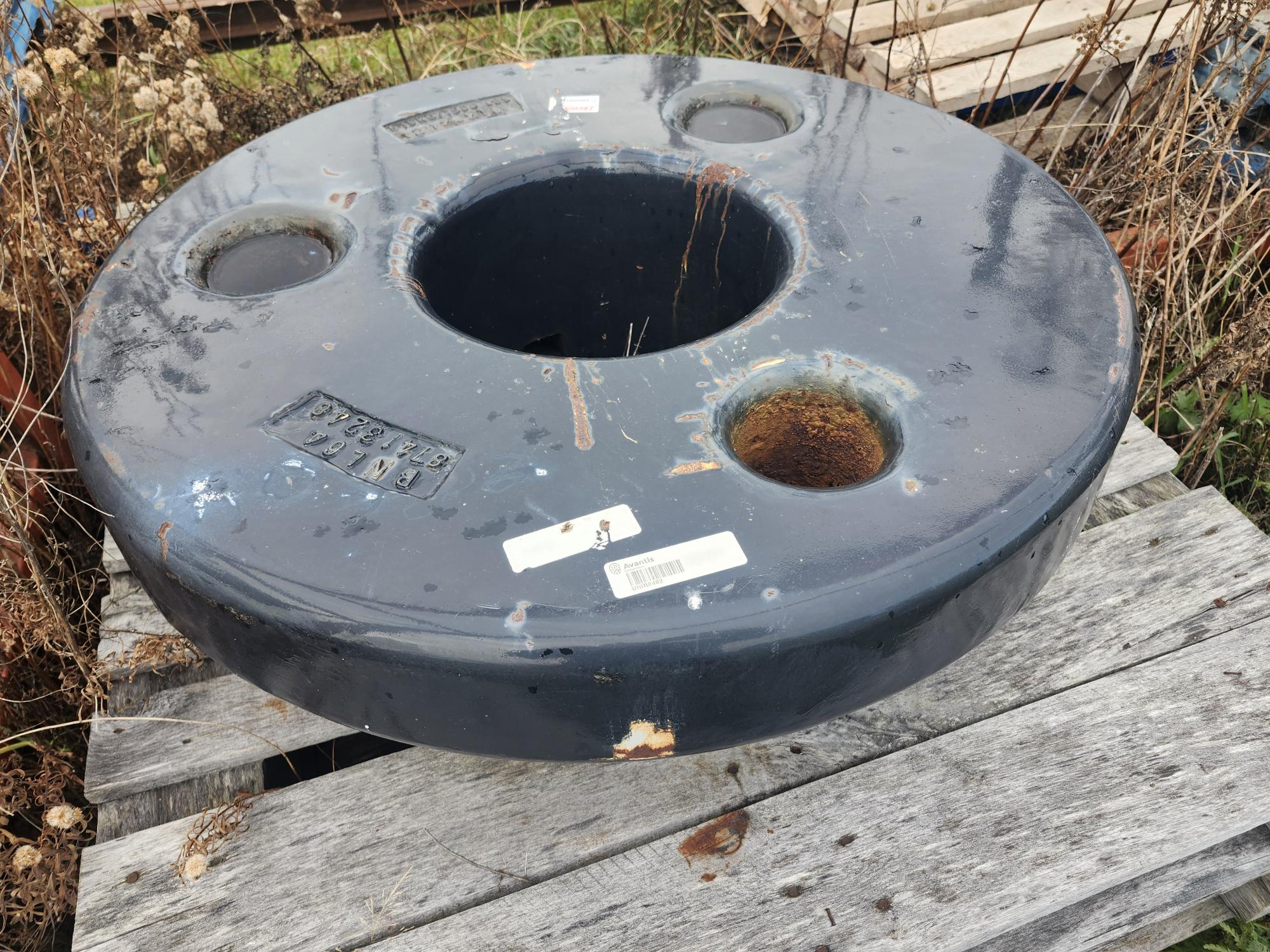 Un grand objet métallique noir, rond et percé de multiples trous repose sur une surface en bois patinée à l'extérieur. De l'herbe sèche entoure la zone et quelques étiquettes et taches de rouille sont visibles sur l'objet.