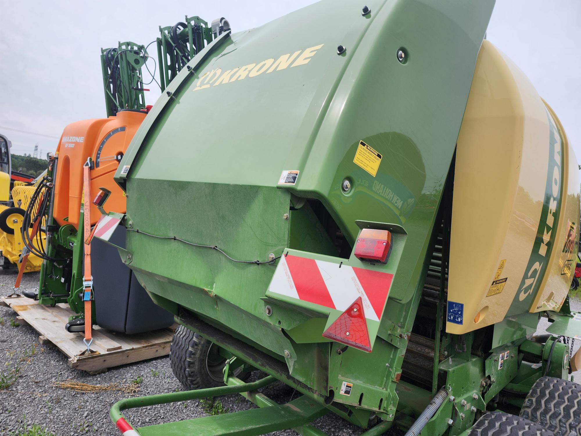 Une machine agricole verte de Krone, probablement une presse à balles, repose sur du gravier à côté d'un équipement orange. La machine est dotée de réflecteurs de sécurité rouges et blancs distinctifs à l'arrière. Plusieurs autres pièces d'équipement agricole sont visibles en arrière-plan.