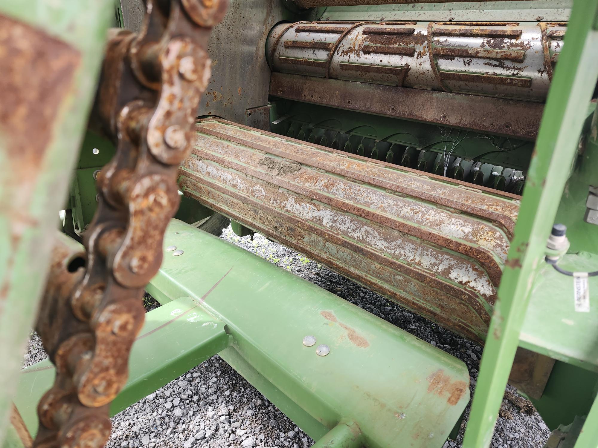 Gros plan d'une machine agricole verte et patinée par les intempéries, avec des composants métalliques rouillés, notamment une grande chaîne et des rouleaux. On peut voir du gravier sur le sol sous la machine.