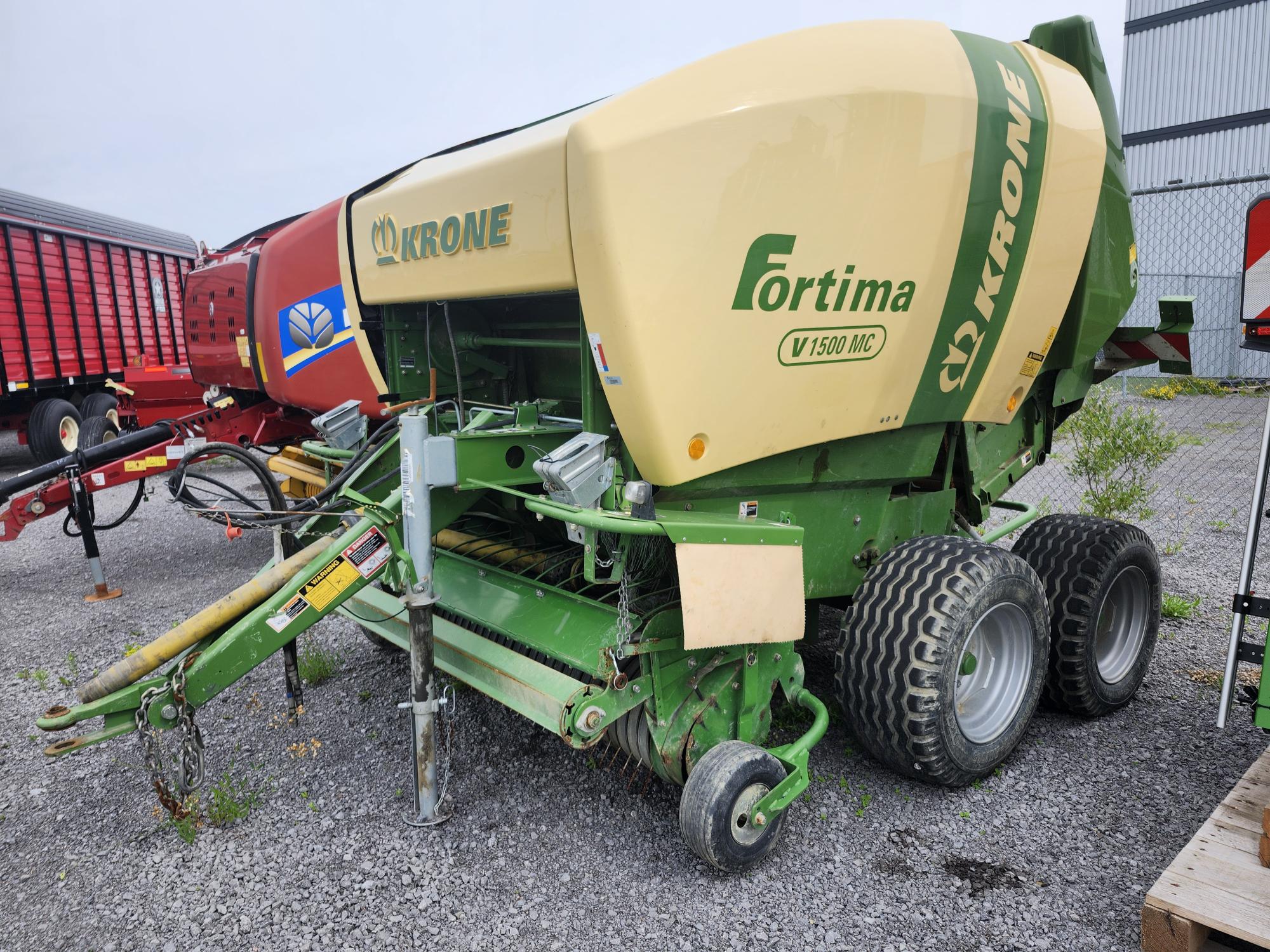 Une presse à balles verte et beige Krone Fortima V1500 MC est garée sur une surface en gravier à l'extérieur. Elle est équipée de grandes roues en caoutchouc et de composants métalliques, avec d'autres machines agricoles visibles en arrière-plan.