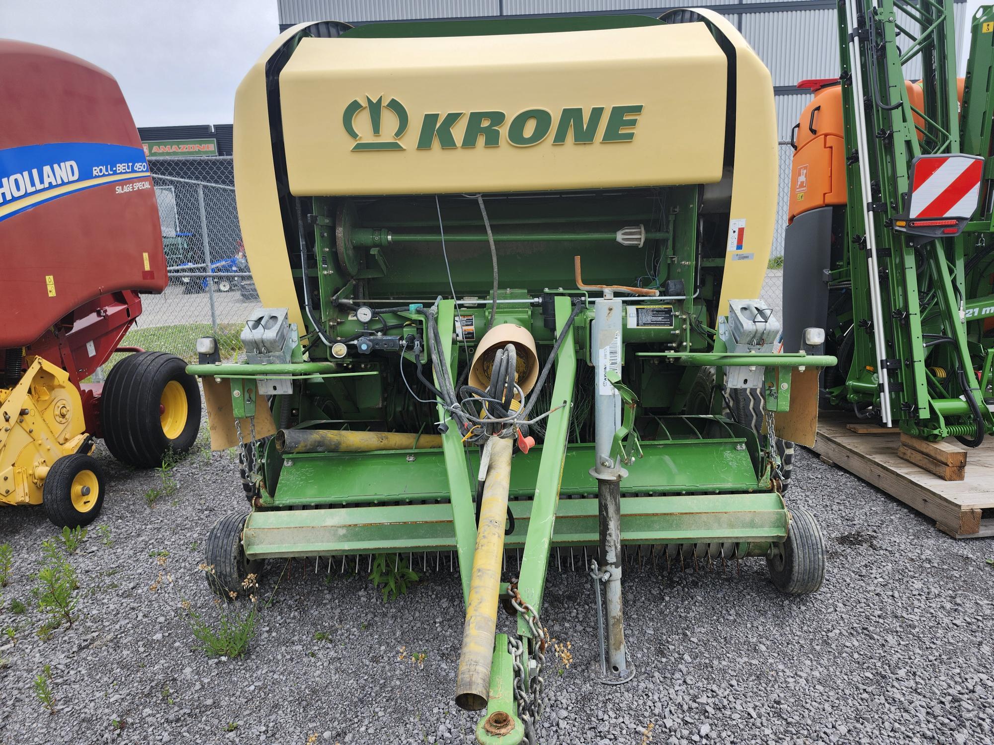 Une machine agricole Krone verte et jaune est posée sur du gravier à l'extérieur, entourée d'autres machines. Ce gros équipement est conçu pour des tâches telles que la mise en balles ou la récolte, avec des composants métalliques robustes et des mécanismes visibles.