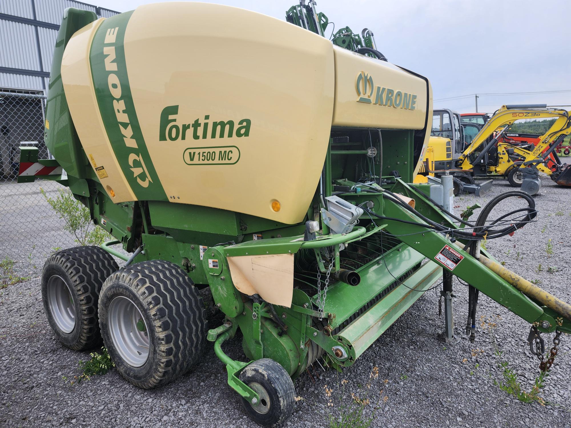 Une presse à balles rondes Krone Fortima V 1500 MC est garée sur du gravier près d'une clôture, avec d'autres machines en arrière-plan. La presse est verte et beige, avec des roues proéminentes et son mécanisme avant visible.