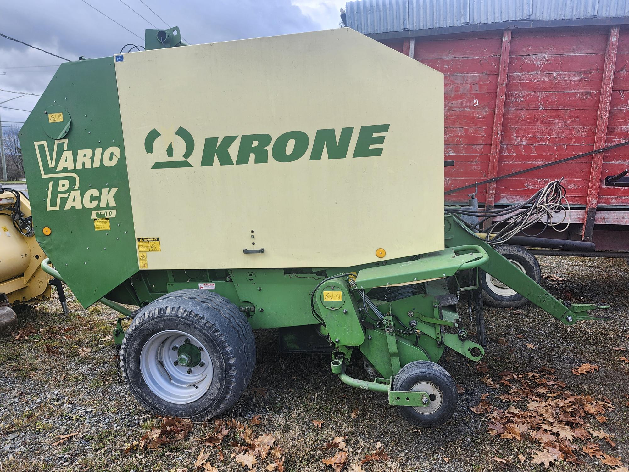 Une presse à balles de foin Krone Vario Pack verte et crème est garée à l'extérieur sur une parcelle d'herbe entourée de feuilles mortes. La presse à balles se trouve à côté d'une structure en bois rouge et d'autres pièces de machines sont visibles en arrière-plan.