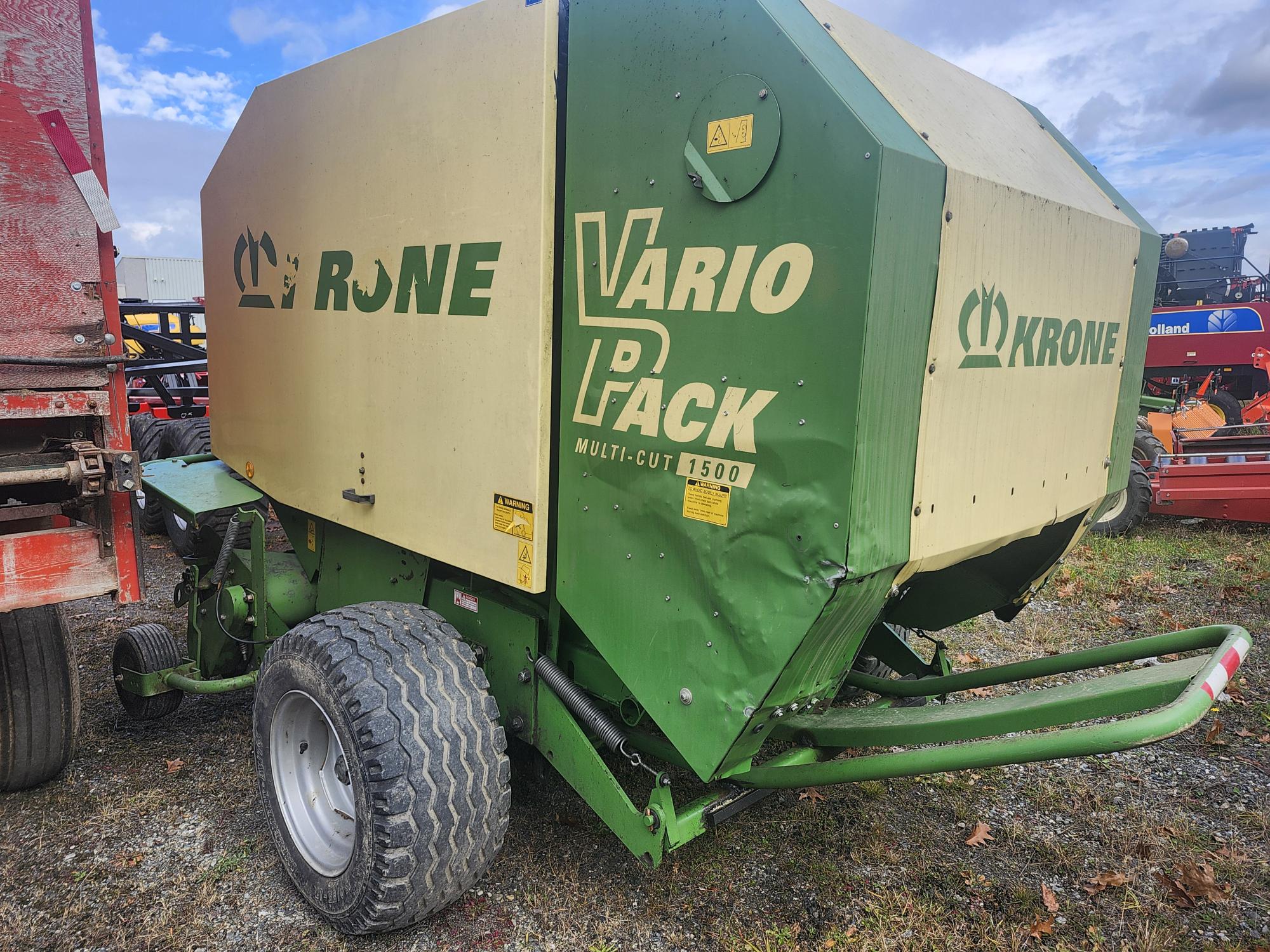 Une presse à balles rondes Krone Vario Pack Multi-Cut 1500 est garée sur une surface en gravier. La machine est verte et beige, avec de gros pneus et un marquage visible sur le côté. On peut voir un ciel nuageux et des parties d'autres équipements en arrière-plan.