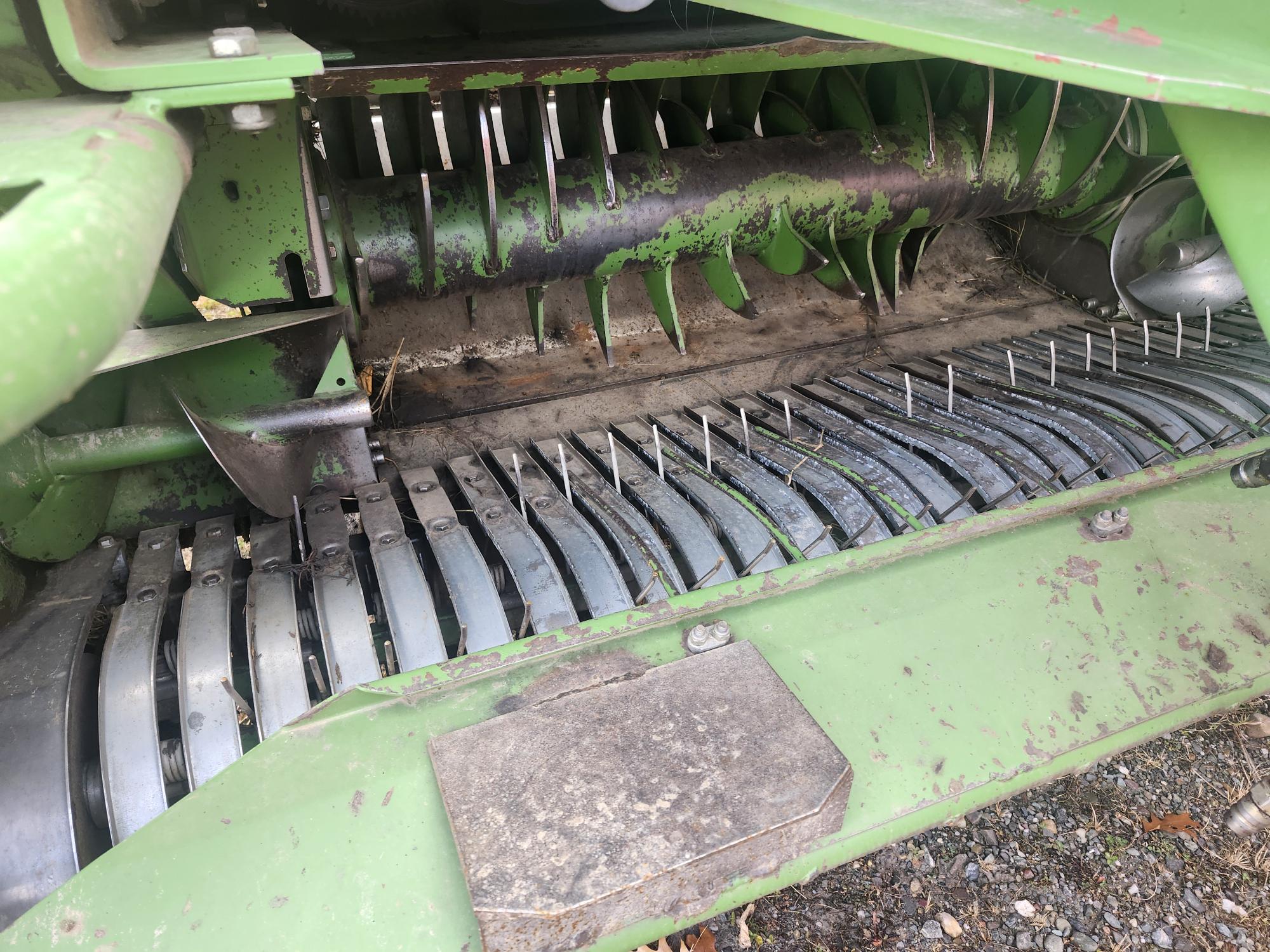 Gros plan d'une pièce de machine agricole verte, avec des lames rotatives et des composants métalliques. La machine présente des signes d'utilisation, avec de l'herbe et des débris accumulés sur les surfaces.