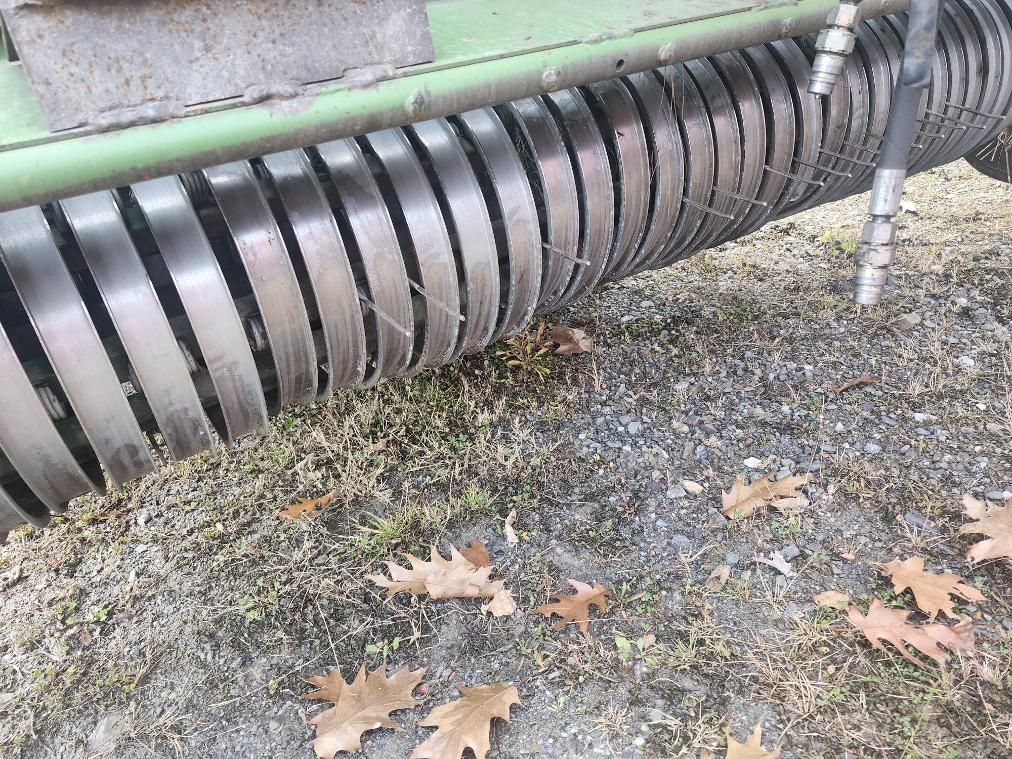 Gros plan sur les rouleaux métalliques d'une machine agricole posés sur le sol, entourés de feuilles sèches et de graviers éparpillés. La machine semble robuste avec une série de disques métalliques parallèles.