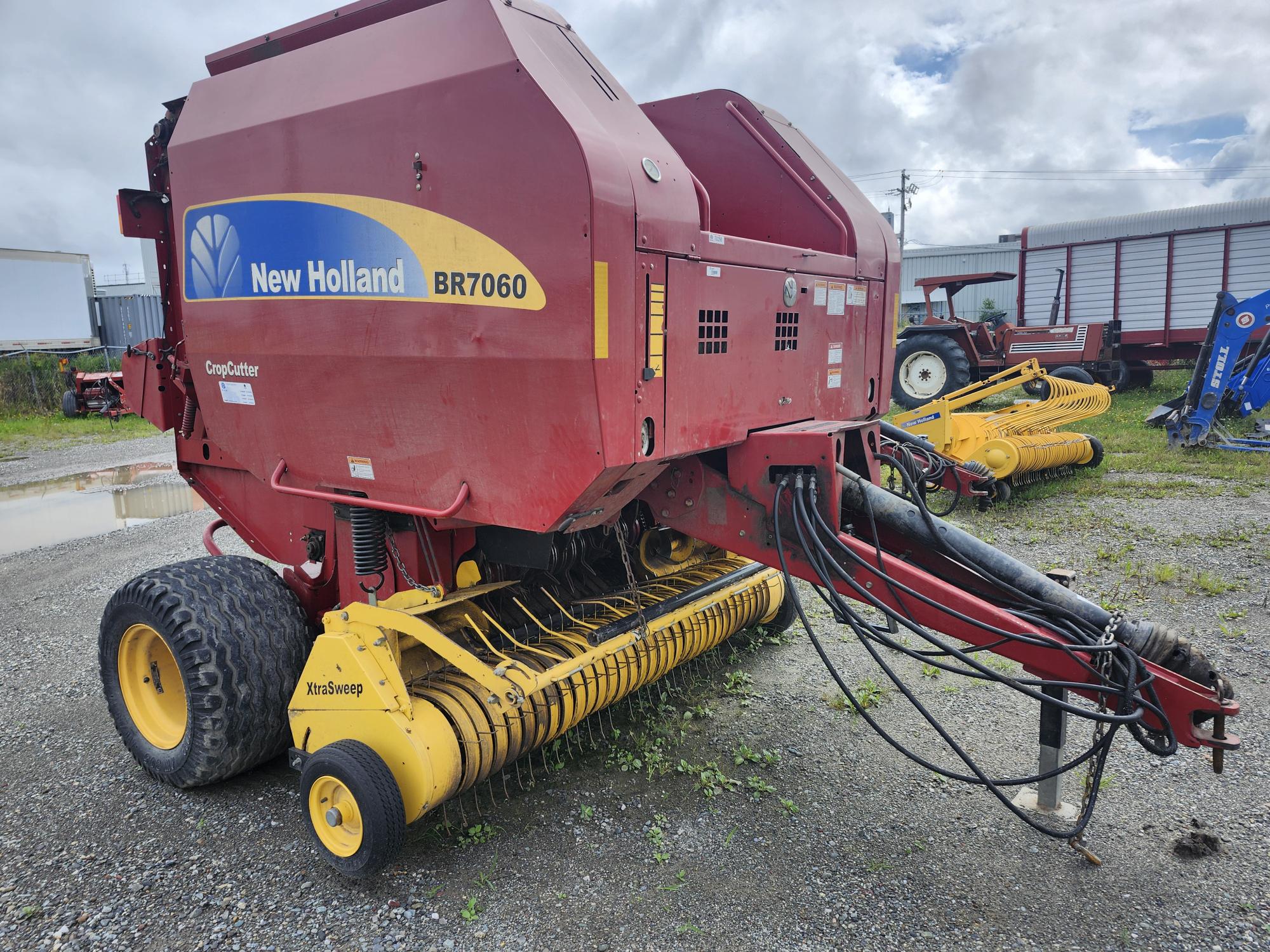 Une presse à balles New Holland BR7060 CropCutter rouge est garée sur du gravier, avec un ramasseur jaune. Elle est entourée d'un ciel nuageux, d'un tracteur et d'équipements agricoles en arrière-plan.