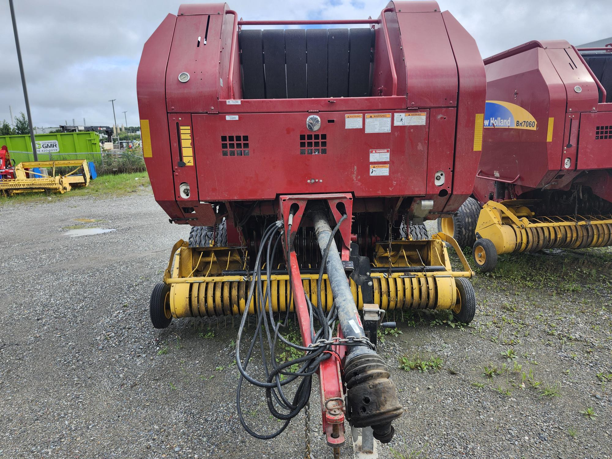 Une machine agricole rouge avec des composants jaunes est garée sur du gravier, probablement une presse à foin, avec diverses pièces mécaniques et tuyaux visibles. À proximité, une autre machine similaire et un peu de verdure sont visibles en arrière-plan.