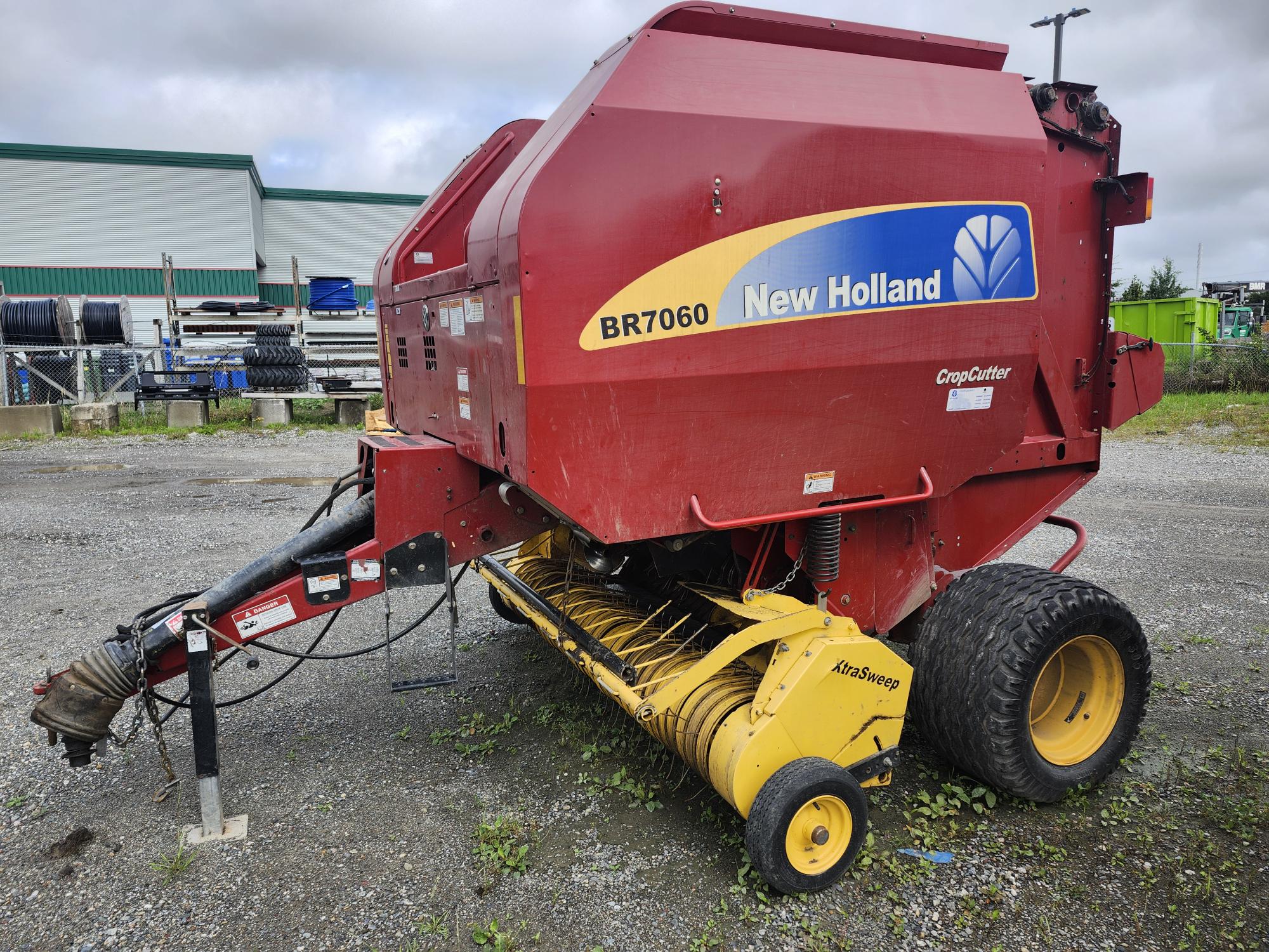 Une presse à balles New Holland BR7060 CropCutter rouge est garée sur une surface en gravier près d'un bâtiment. La presse à balles présente des détails jaunes, de grandes roues et est équipée pour une utilisation agricole. Des piles de pneus et de machines sont visibles en arrière-plan.