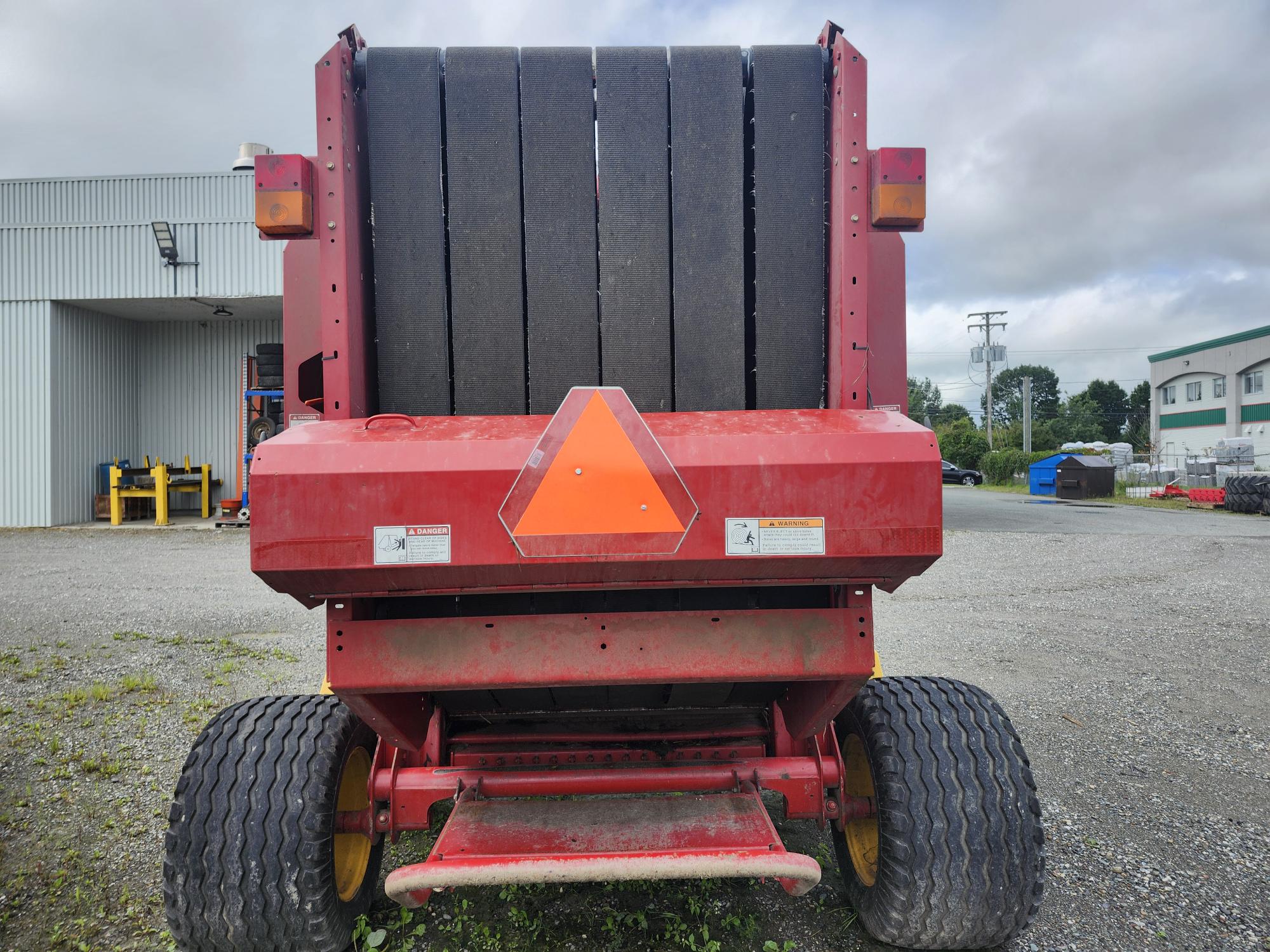 Une presse agricole rouge avec un panneau réfléchissant triangulaire à l'arrière est garée sur une surface en gravier près d'un bâtiment métallique. Le ciel est nuageux et il y a d'autres bâtiments et machines en arrière-plan.