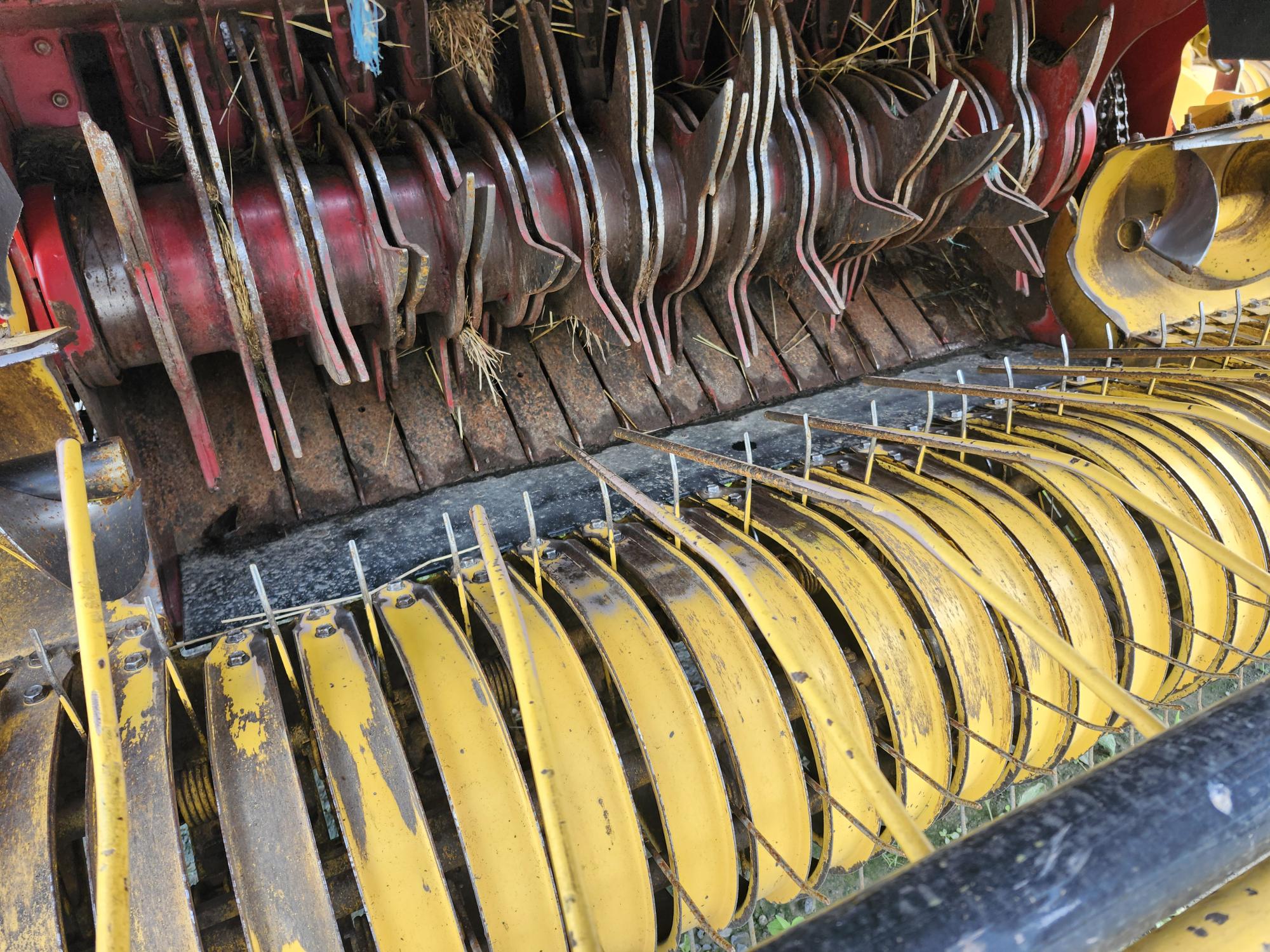 Gros plan d'une machine agricole jaune et rouge, montrant ses dents et ses lames rotatives, conçues pour la récolte des cultures. L'équipement est usé par les intempéries, avec du foin et des débris coincés dans le mécanisme.