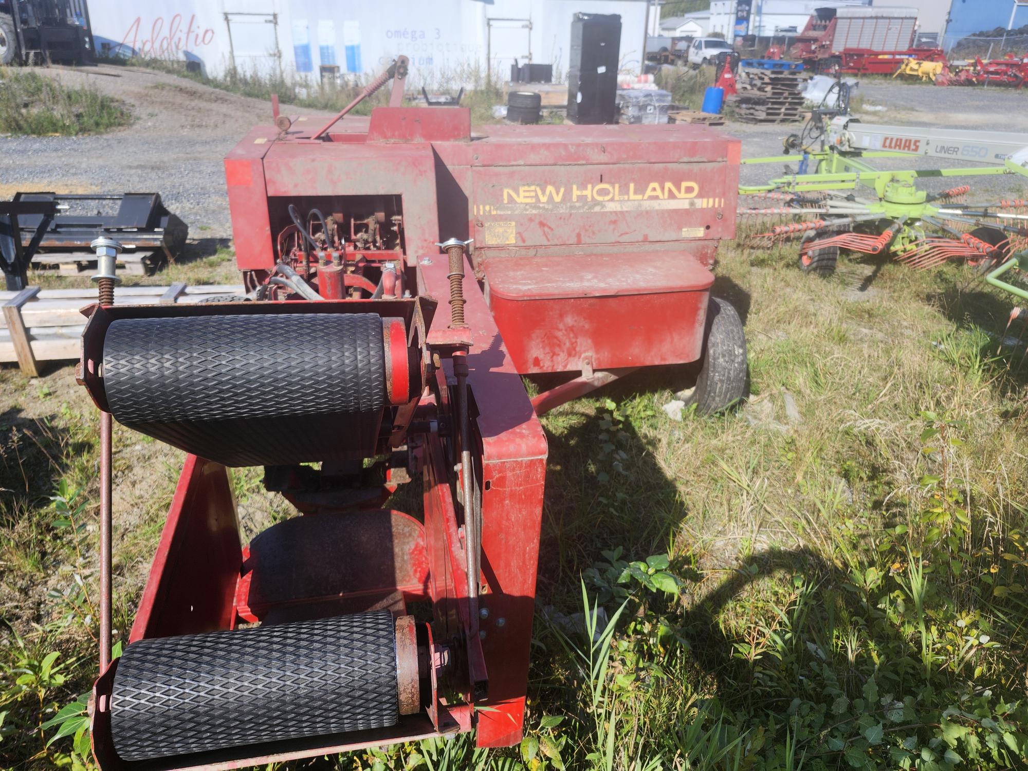 Une machine agricole New Holland rouge est garée dans une zone herbeuse à proximité de bâtiments industriels et d'autres équipements agricoles. La machine présente des signes d'utilisation et de vieillissement, avec des pièces usées et de la rouille visibles. Les ombres et la lumière du soleil mettent en valeur sa structure.