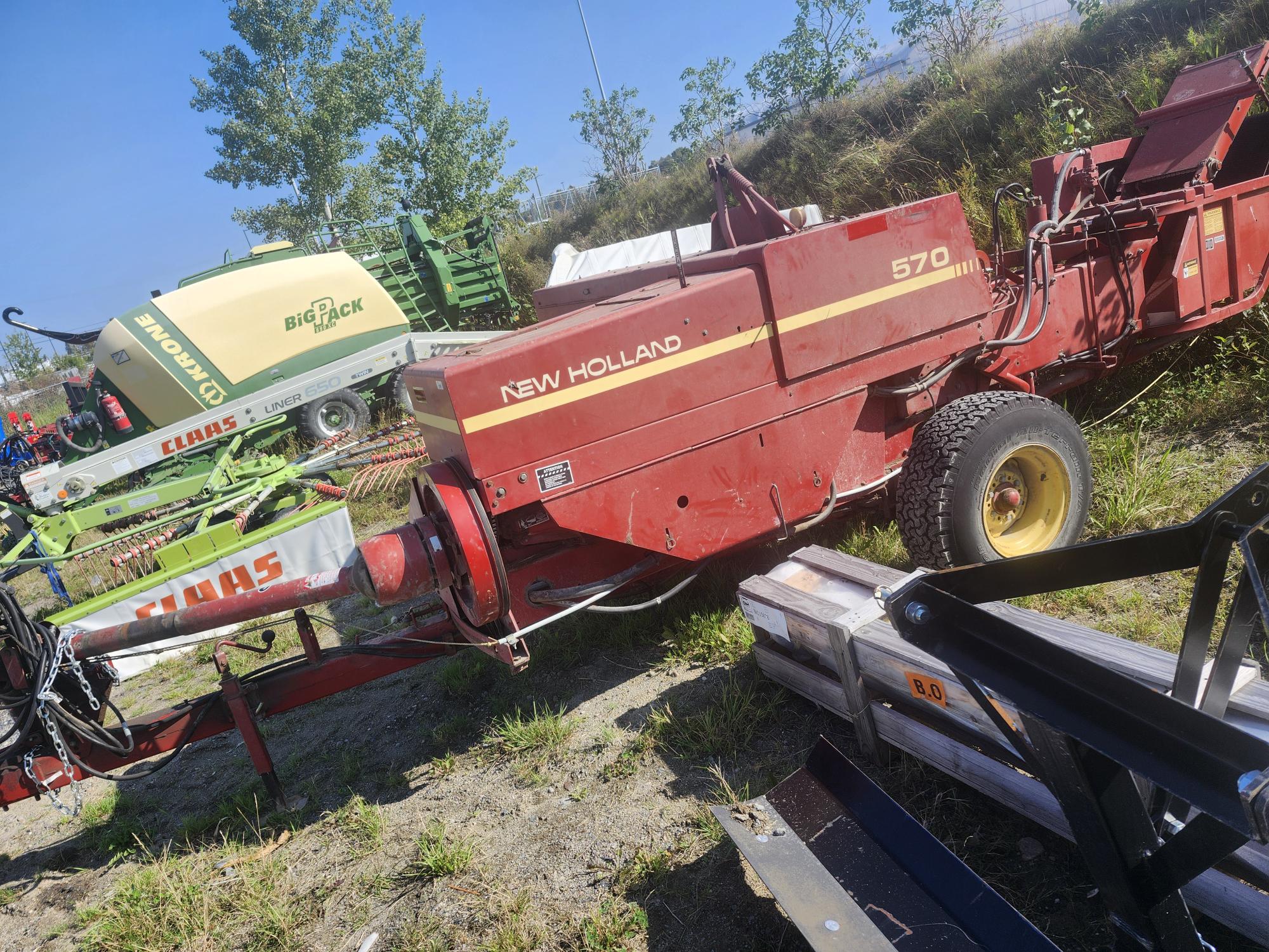 Une presse à balles de foin New Holland 570 d'époque est garée sur l'herbe, entourée de diverses machines agricoles. La presse a une carrosserie rouge avec des roues jaunes. À l'arrière-plan, on voit des machines Claas vertes et jaunes. Des arbres bordent la zone derrière.