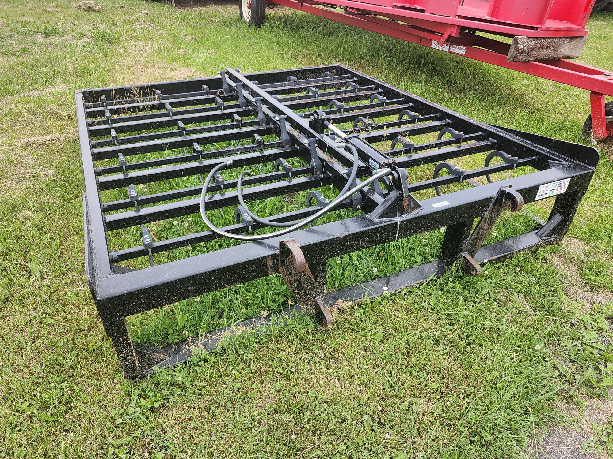 Un accessoire agricole en métal noir avec plusieurs barres et crochets est posé sur l'herbe verte. Il est doté de composants hydrauliques et se trouve à côté d'une machine agricole rouge.