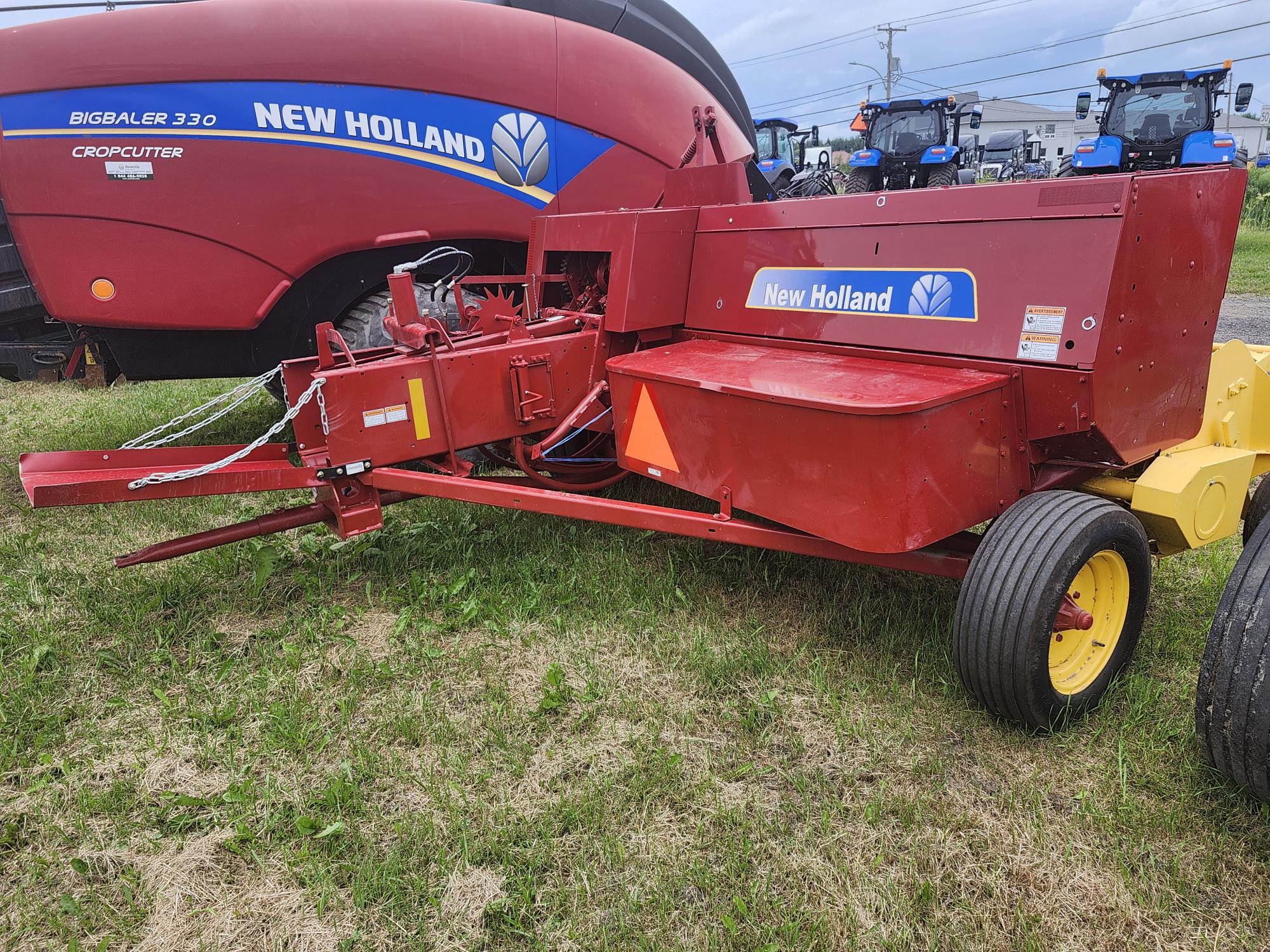 Une faucheuse New Holland rouge est posée sur l'herbe verte. Elle est attachée à une remorque à roues jaunes. Une presse à balles rouge plus grande avec le logo New Holland est visible à l'arrière-plan, à côté de tracteurs bleus sous un ciel nuageux.