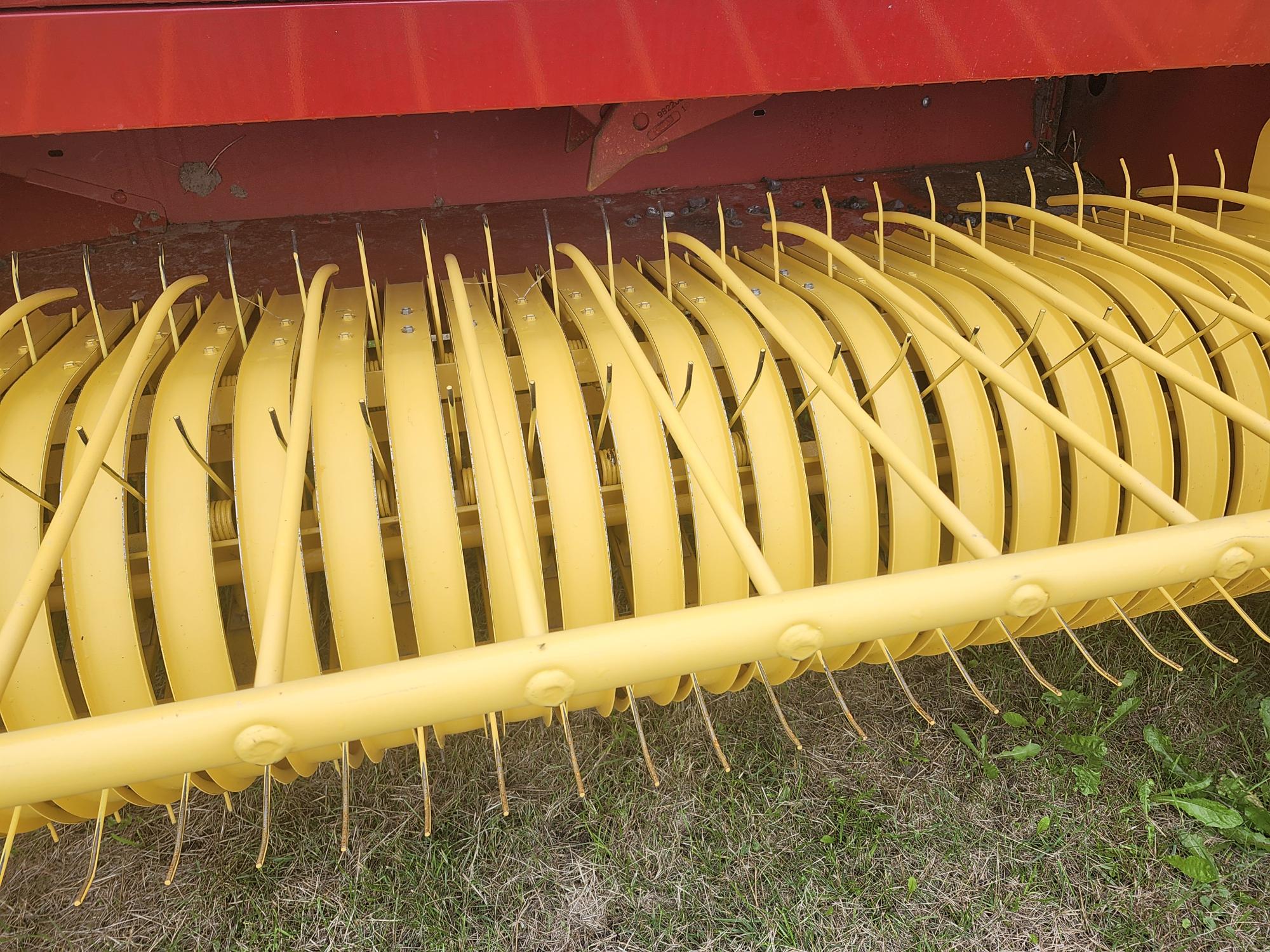 Gros plan d'une pièce de machine agricole jaune avec des dents courbées, probablement une partie d'un râteau à foin ou d'une presse à balles, sur un fond d'herbe verte et d'un cadre en métal rouge.