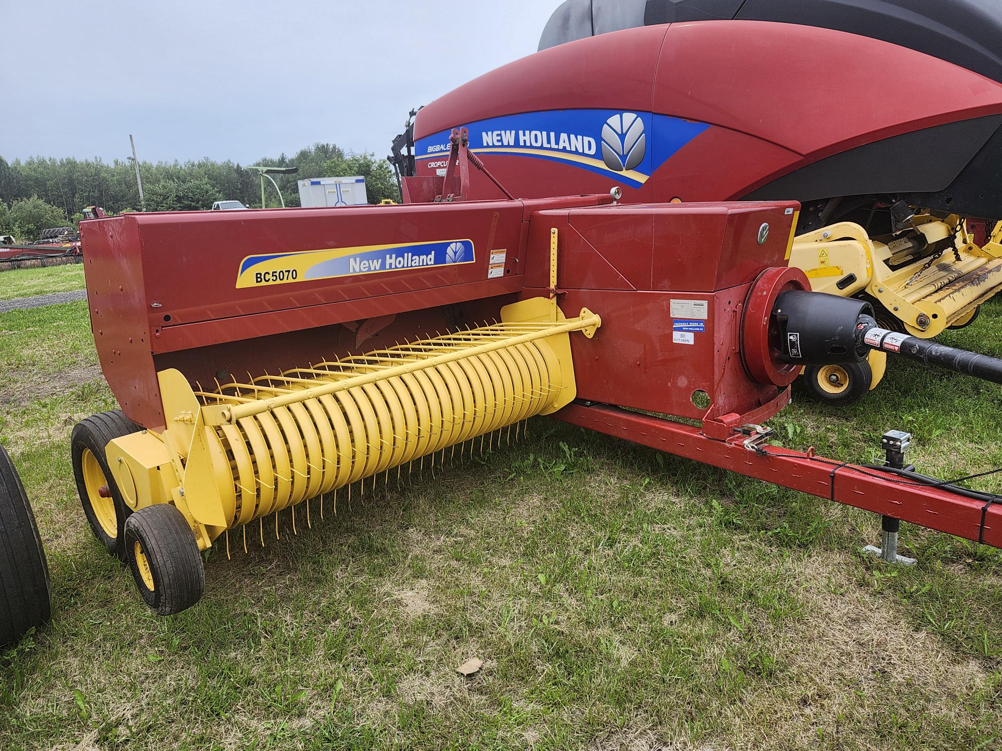 Une presse à balles rouge et jaune New Holland BC5070 est garée sur l'herbe. La presse à balles est équipée d'un grand tambour de ramassage jaune et d'un corps cylindrique. Un tracteur est partiellement visible en arrière-plan, avec des arbres au loin.