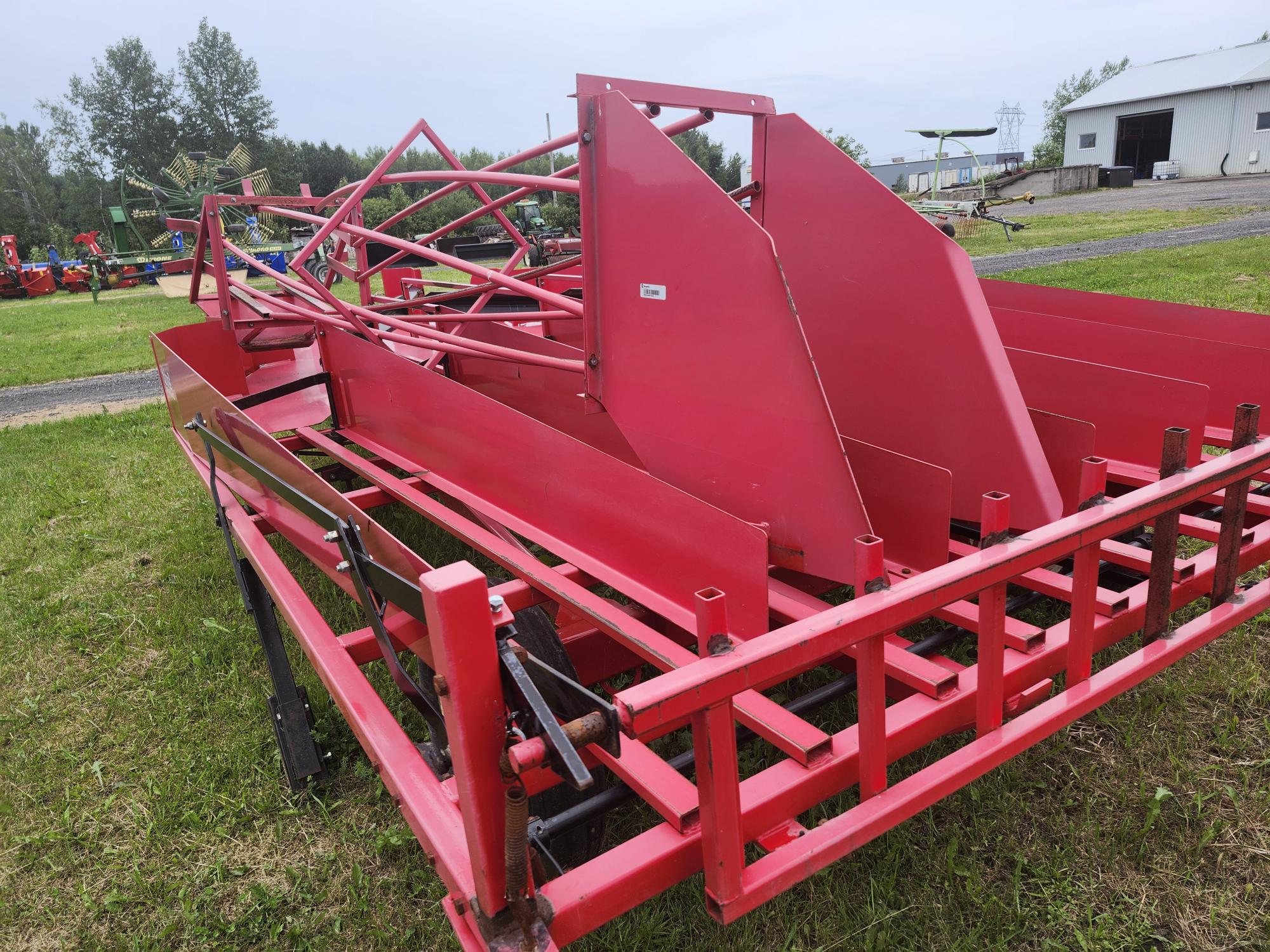 Une machine agricole rouge avec divers composants métalliques et un cadre à lattes est positionnée sur de l'herbe verte. Un bâtiment et des arbres sont visibles en arrière-plan sous un ciel nuageux.