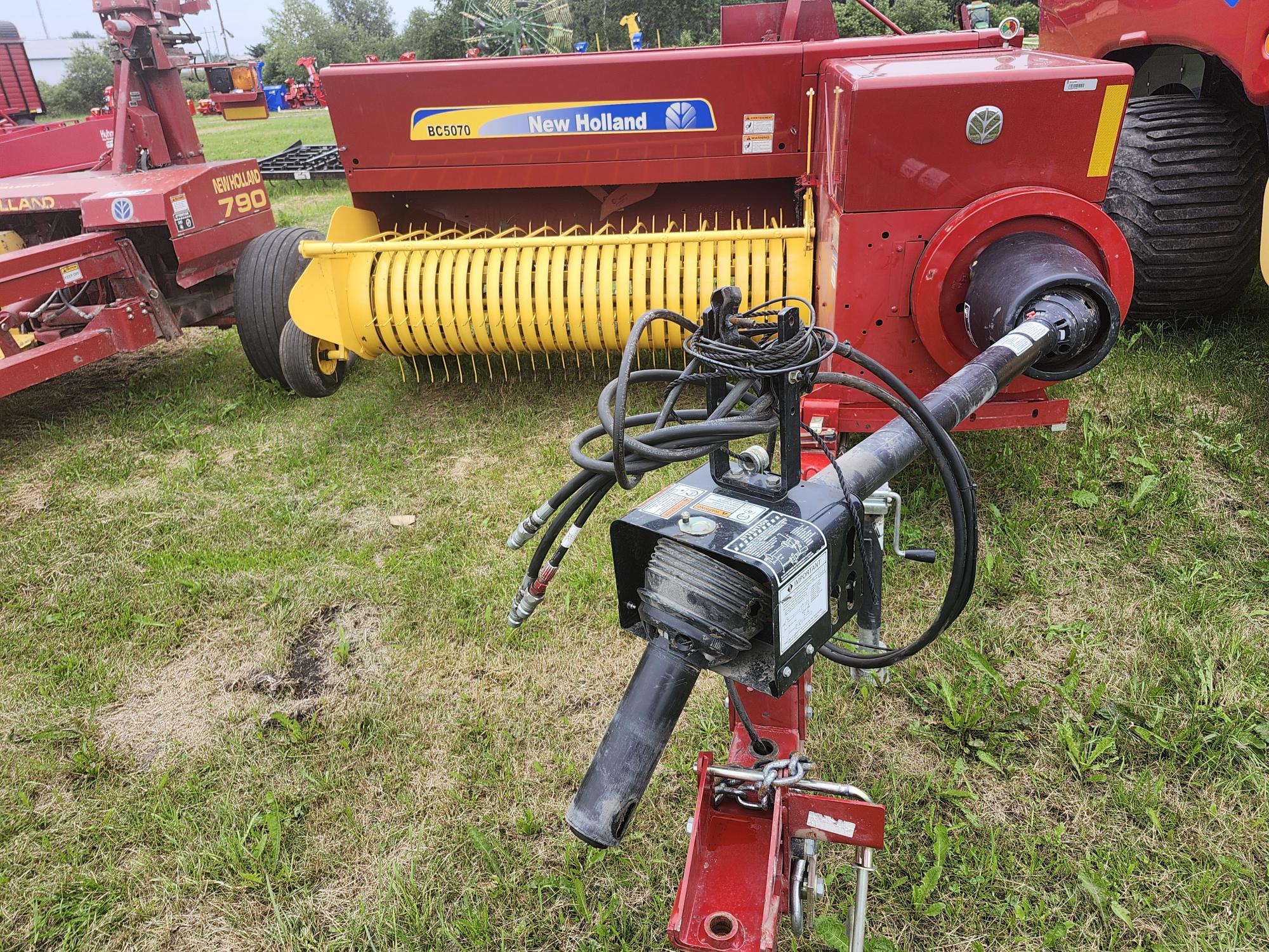 Une presse à foin rouge et jaune New Holland portant le numéro de modèle BC5070 est garée sur l'herbe. Elle est dotée de plusieurs composants, notamment des tuyaux hydrauliques et des engrenages, sur fond d'autres équipements agricoles et d'un champ herbeux.