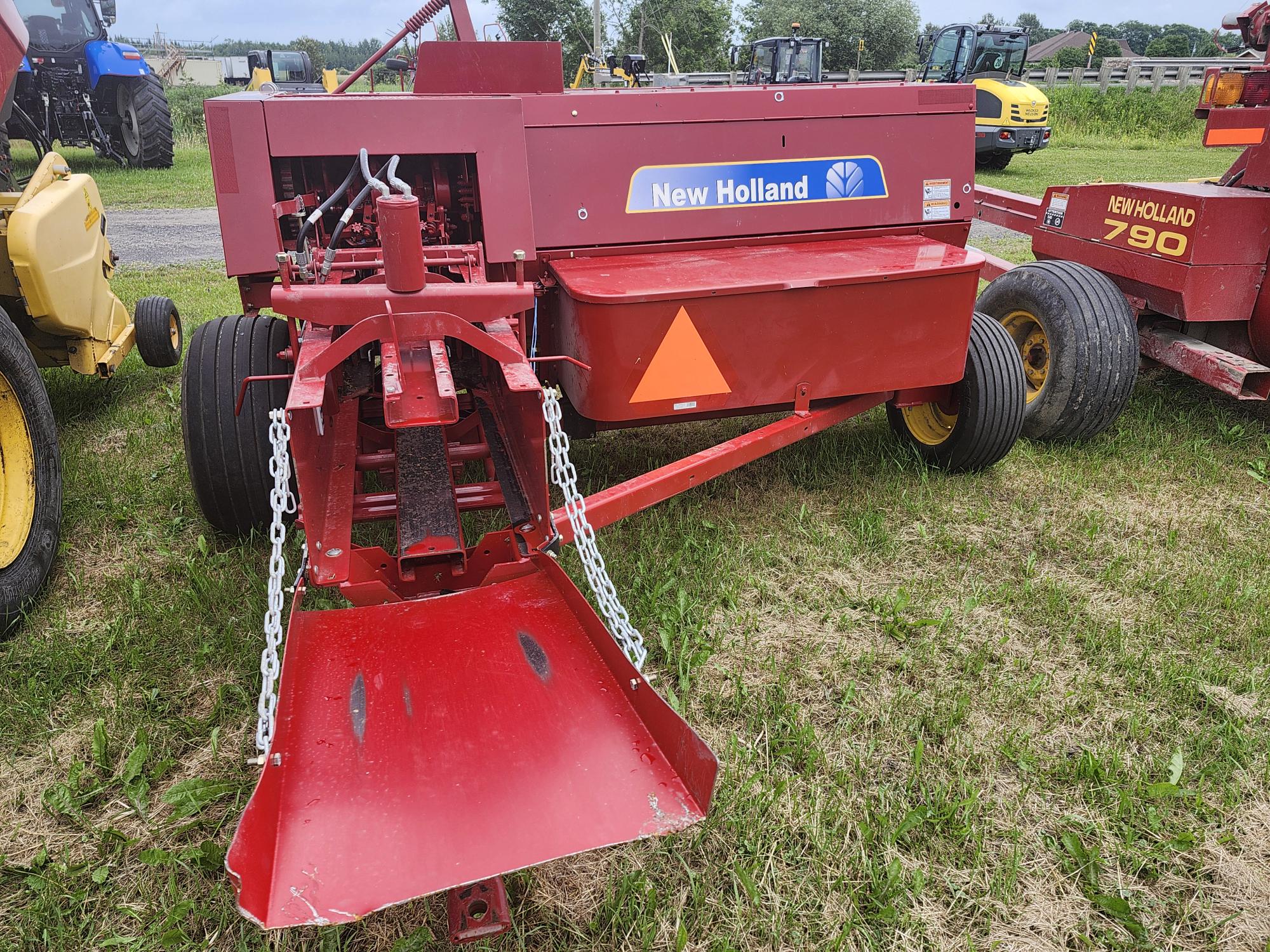 Une presse agricole New Holland rouge équipée d'une grande goulotte et d'accessoires à chaîne est exposée sur un terrain herbeux. D'autres machines sont visibles à l'arrière-plan.