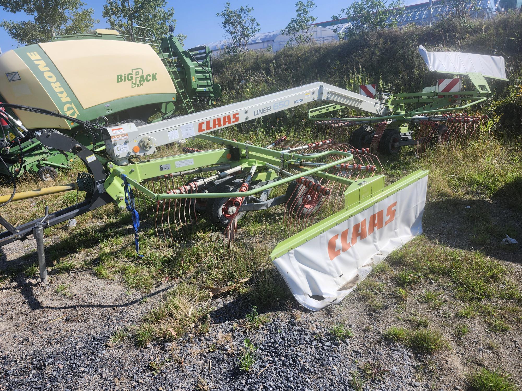 Un andaineur double Claas Liner 650 vert est stationné sur une pelouse avec une presse à balles Krone Big Pack à proximité. L'équipement est entouré d'herbe clairsemée et de graviers épars sous un ciel bleu clair.