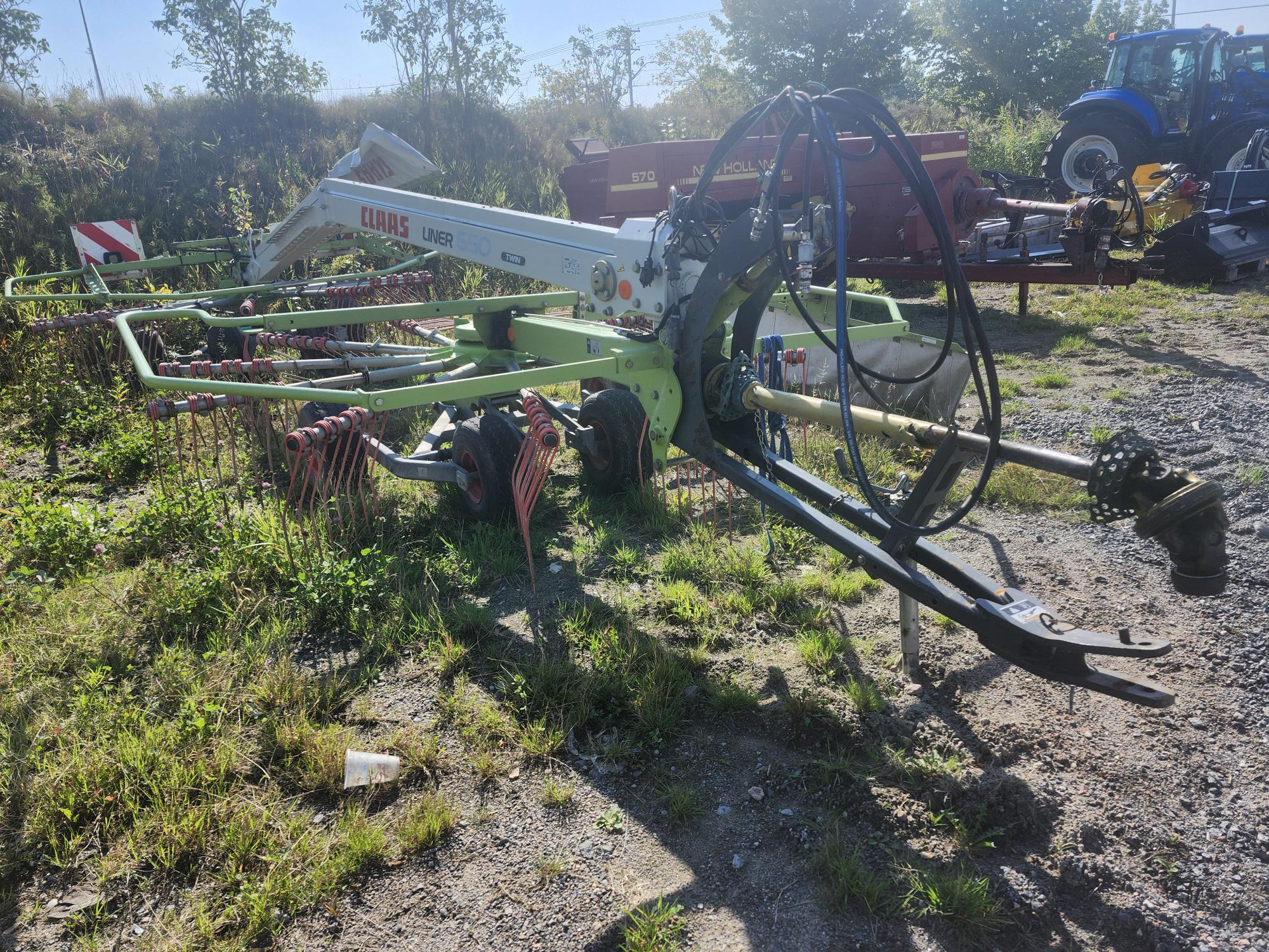 Une machine agricole, probablement un râteau à foin, est garée sur une zone herbeuse et gravillonnée. Elle est dotée de plusieurs gros éléments en forme de roues et de bras métalliques. À l'arrière-plan, il y a des arbres, d'autres machines et un tracteur bleu. Le ciel est clair et ensoleillé.