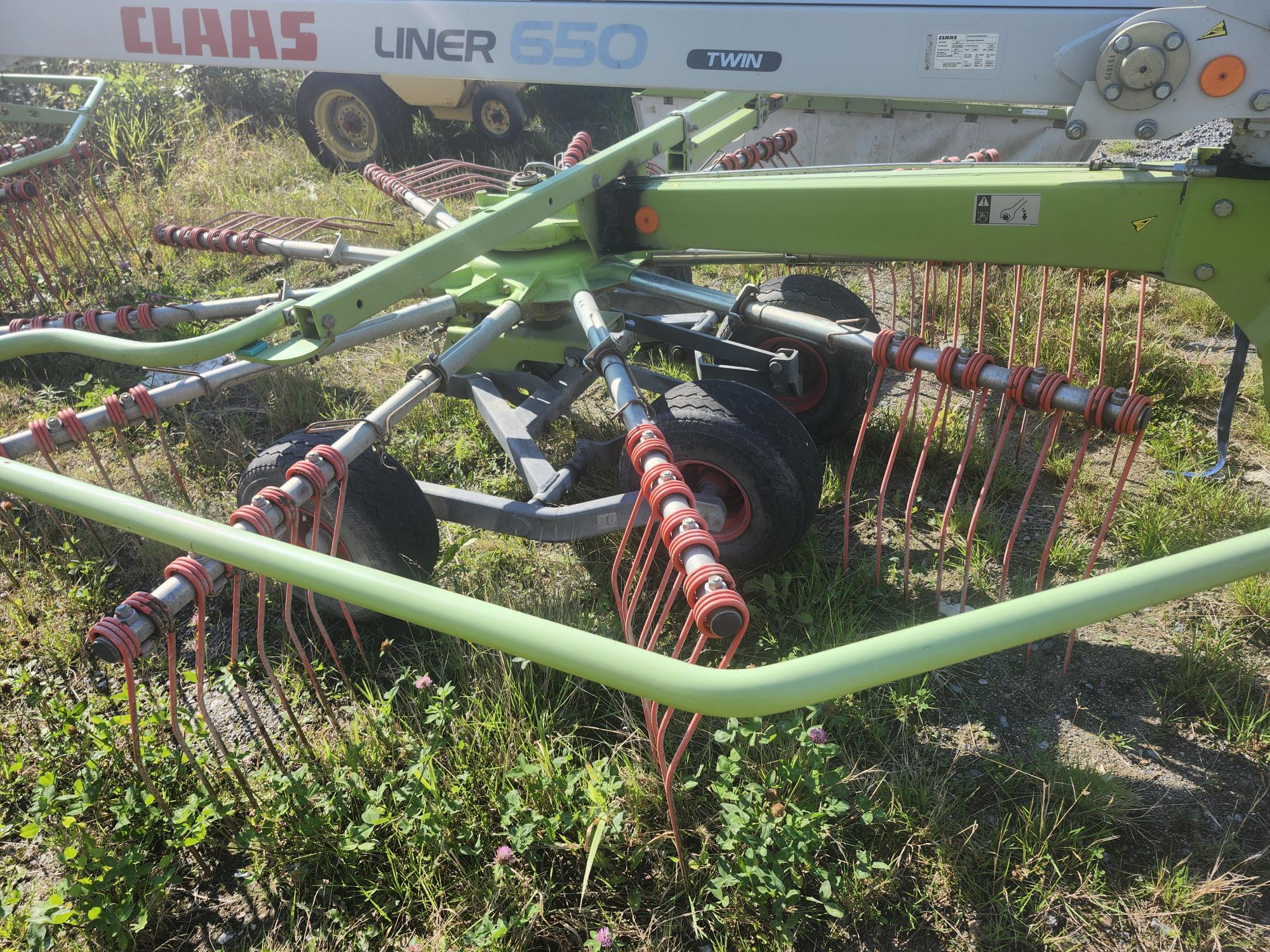 Gros plan d'une machine agricole équipée de plusieurs râteaux pour récolter les récoltes, installée dans une zone herbeuse. La machine est étiquetée « CLAAS LINER 650 TWIN » et comporte des composants verts et oranges avec des bobines et des roues en métal.