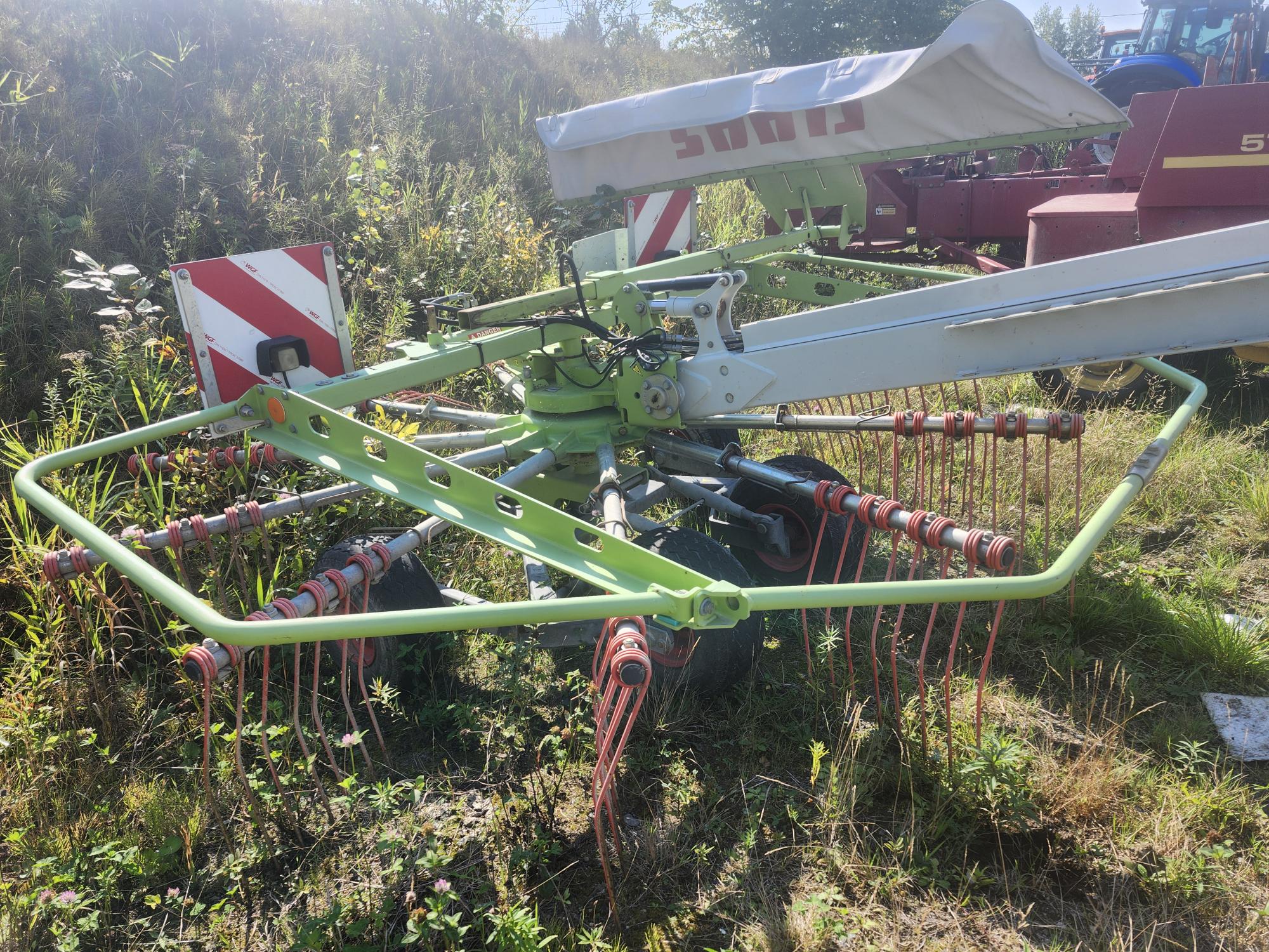 Une machine agricole verte à râteau rotatif avec plusieurs bras métalliques et des dents rouges, posée sur l'herbe avec d'autres équipements agricoles en arrière-plan, sous la lumière du soleil.