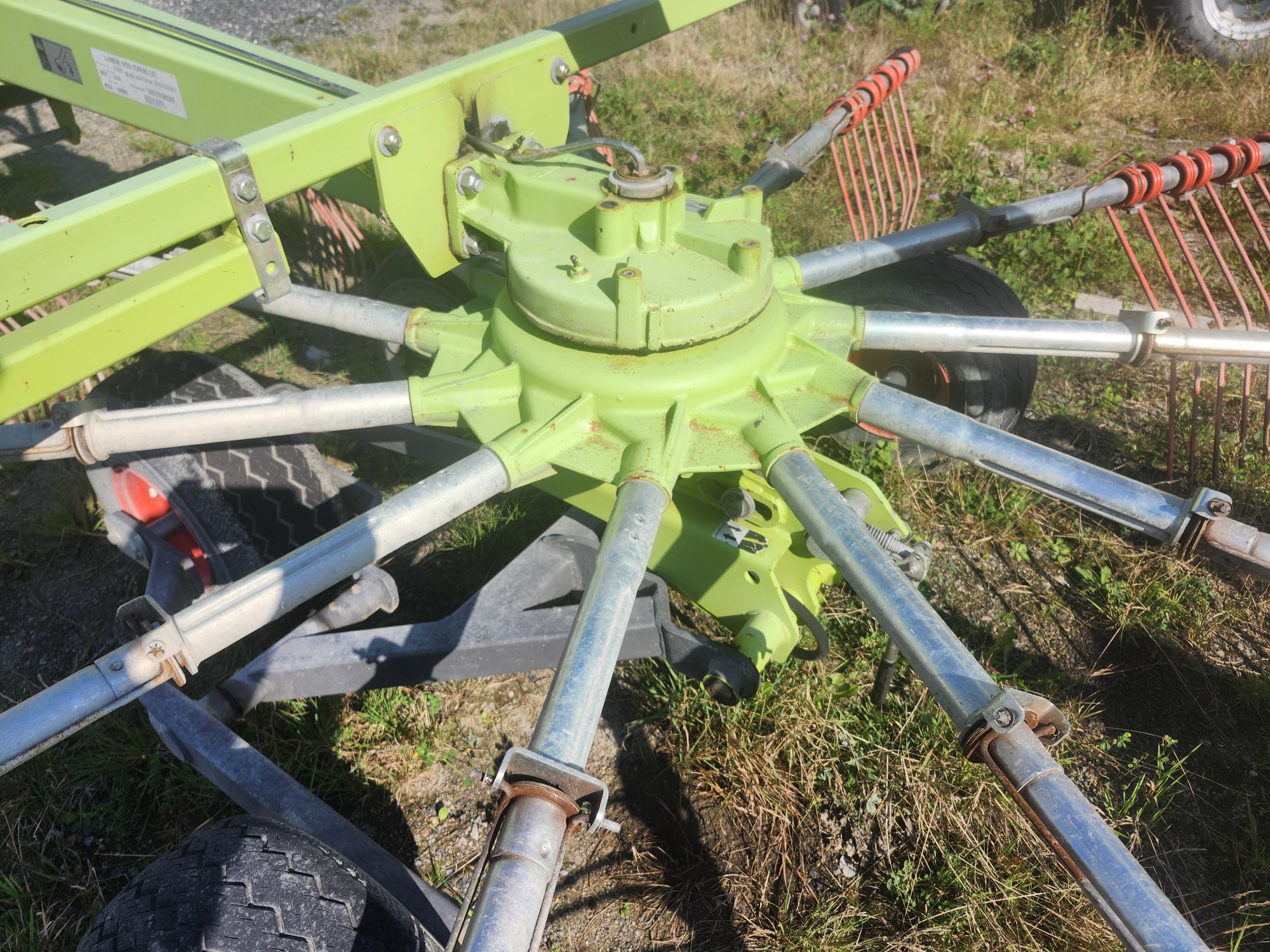 Gros plan d'une machine agricole verte dotée de plusieurs bras métalliques et d'accessoires en forme de peigne rouge, posée sur une pelouse rugueuse avec une zone de gravier à proximité. Les roues de la machine et une partie du châssis sont visibles.