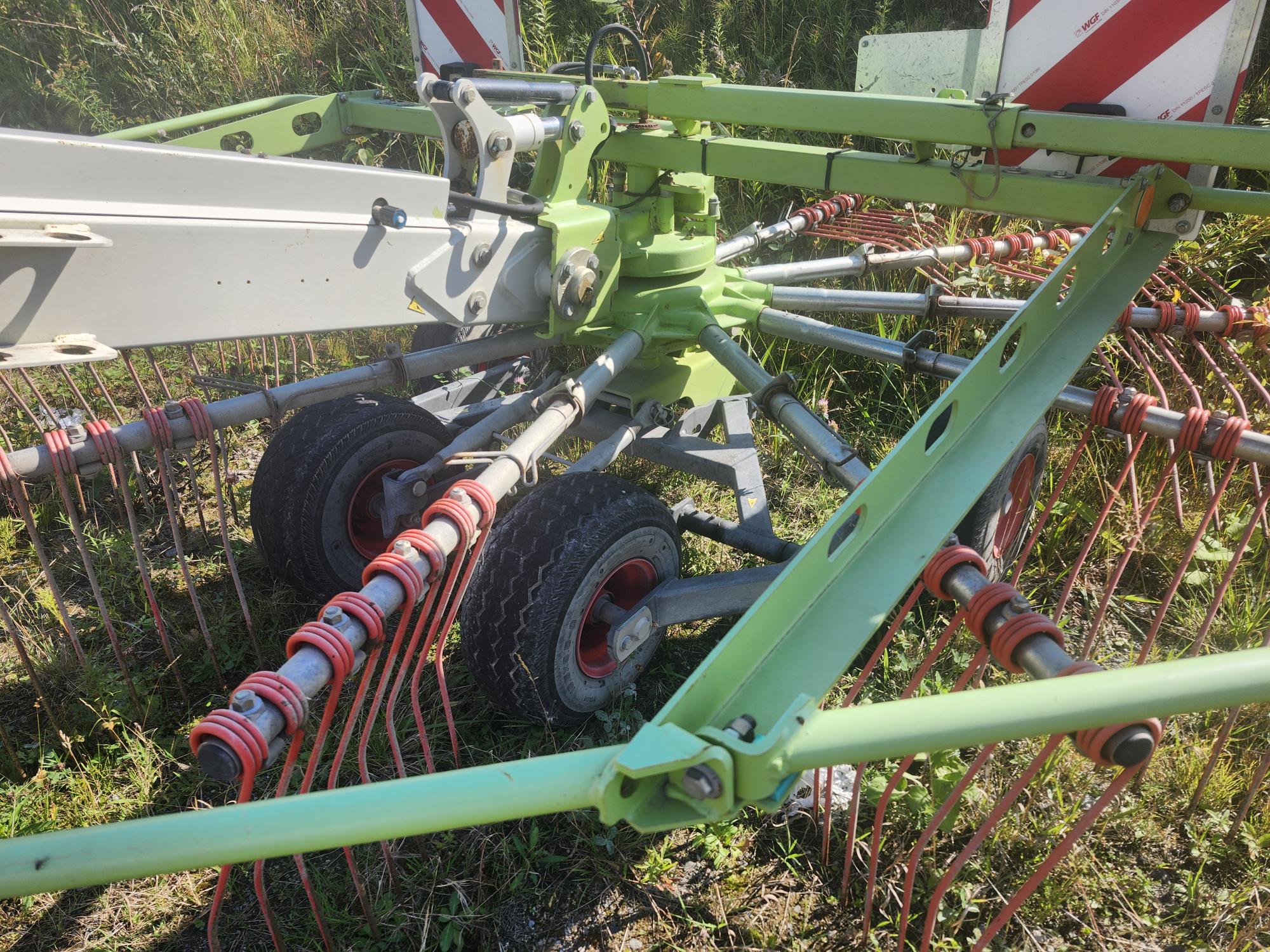 Gros plan d'une machine agricole à dents et roues multiples, utilisée pour épandre de l'herbe ou du foin. L'équipement est situé dans une zone herbeuse, avec des pièces métalliques vert vif et des accents rouges.