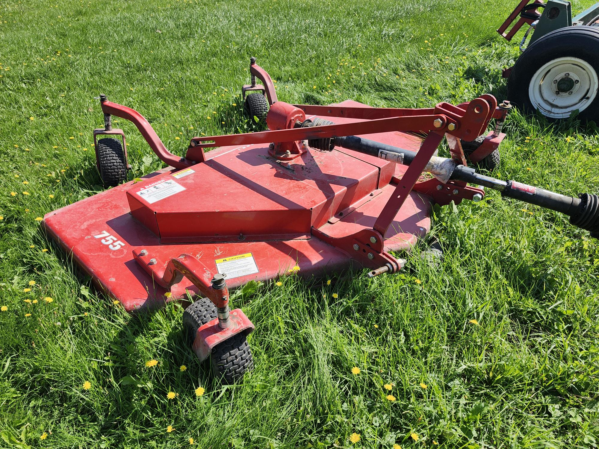 Une tondeuse rotative rouge à plusieurs roues est posée sur une pelouse verte luxuriante, prête à être attelée à un tracteur en arrière-plan. Des pissenlits jaune vif sont dispersés sur l'herbe.